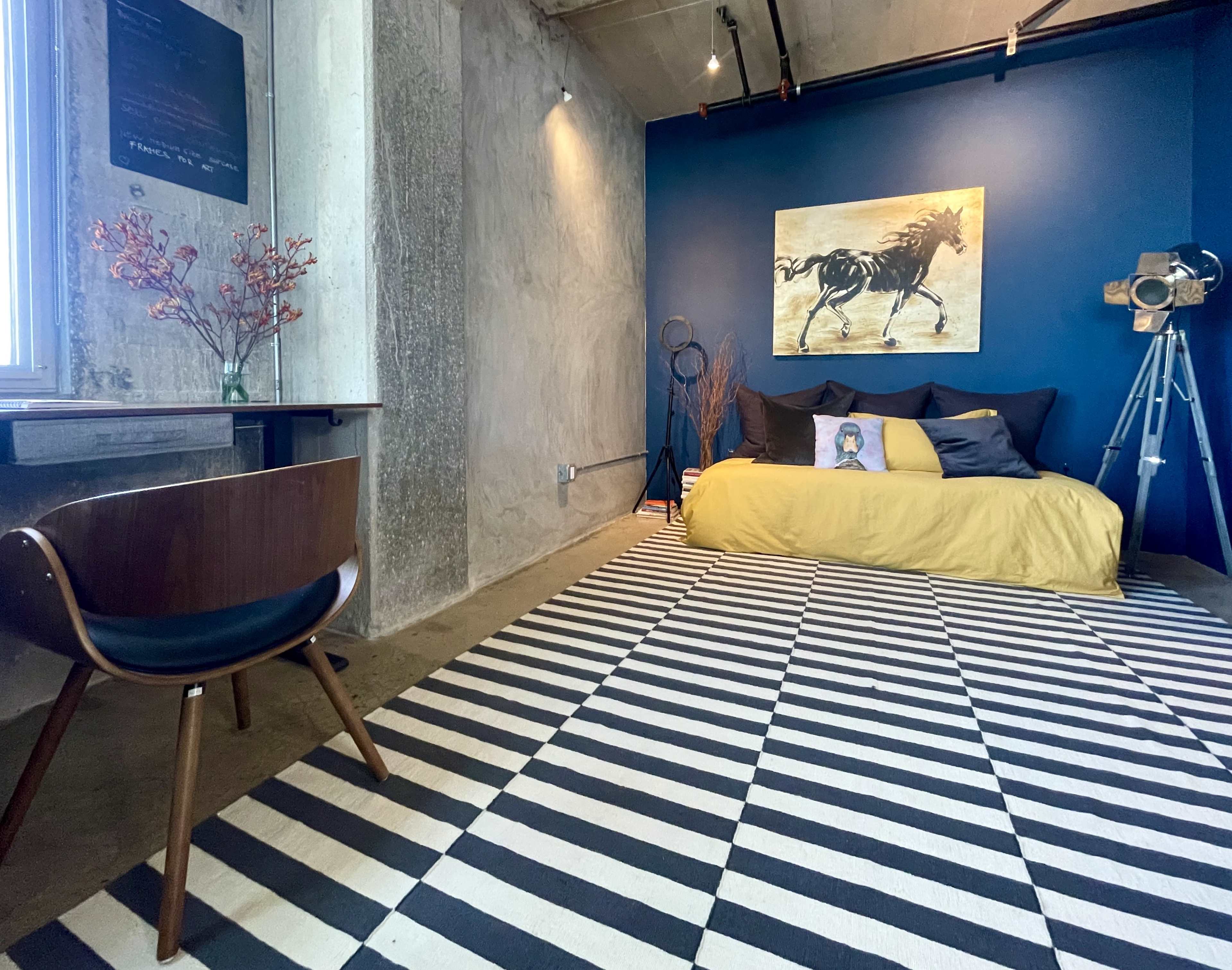 The image shows a modern room featuring a yellow bedspread on a low bed, a wooden chair, a large wall art of a horse, and a striped rug.