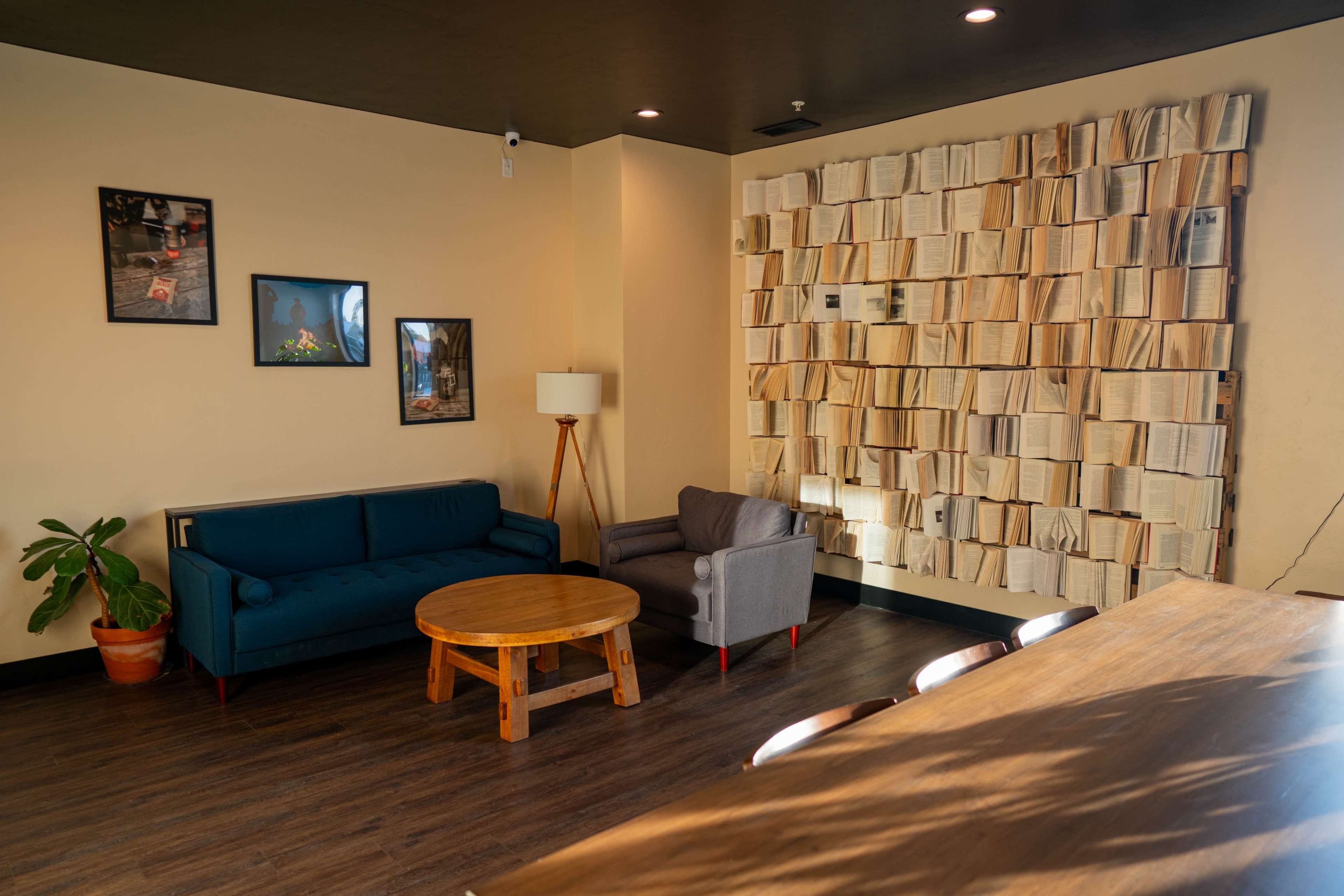The image shows a cozy seating area with a blue couch, a gray armchair, a wooden round coffee table, and a wall built from stacked books.