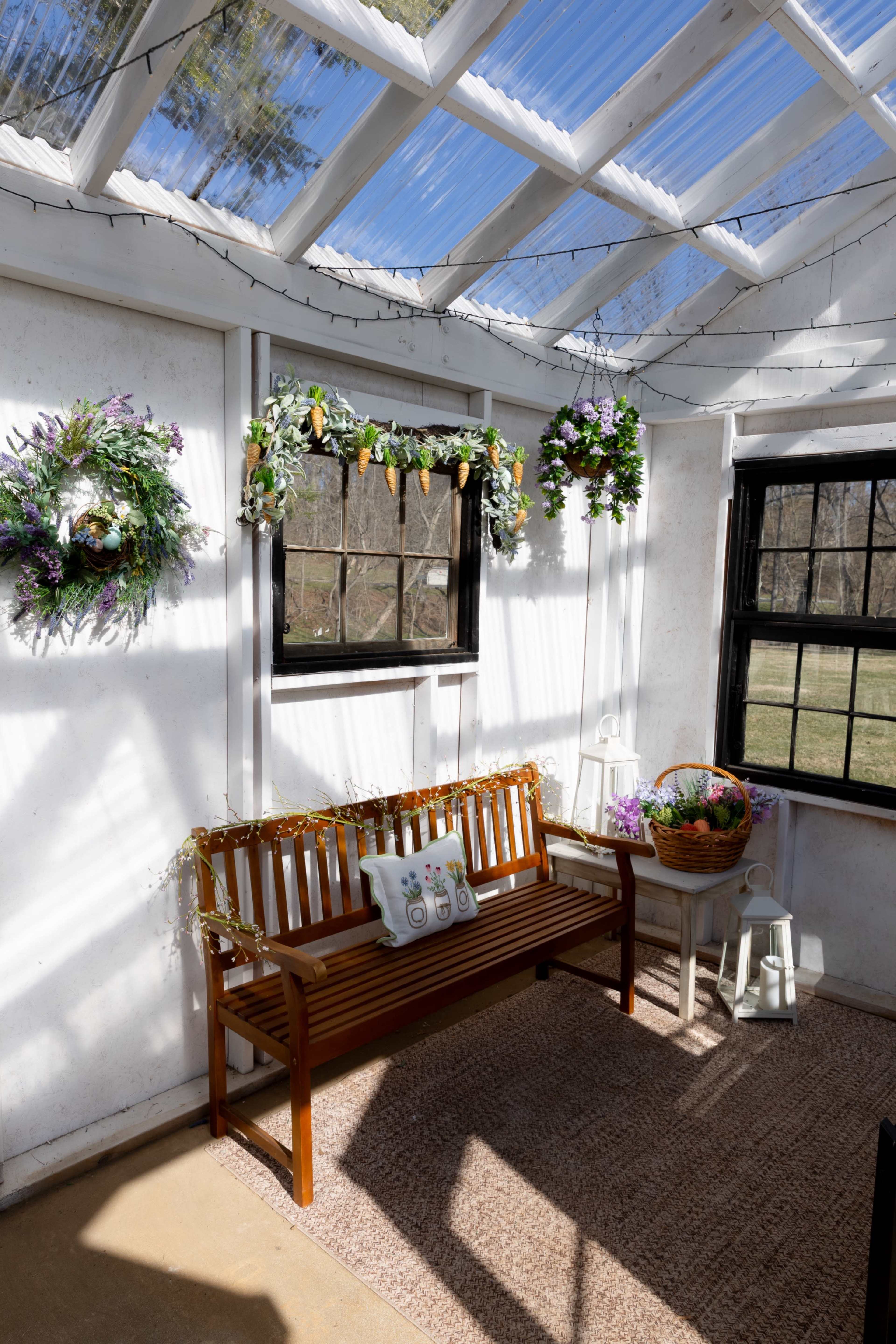 The interior of a sunlit greenhouse features a wooden bench, decorative wreaths hanging on the walls, and a basket filled with flowers.