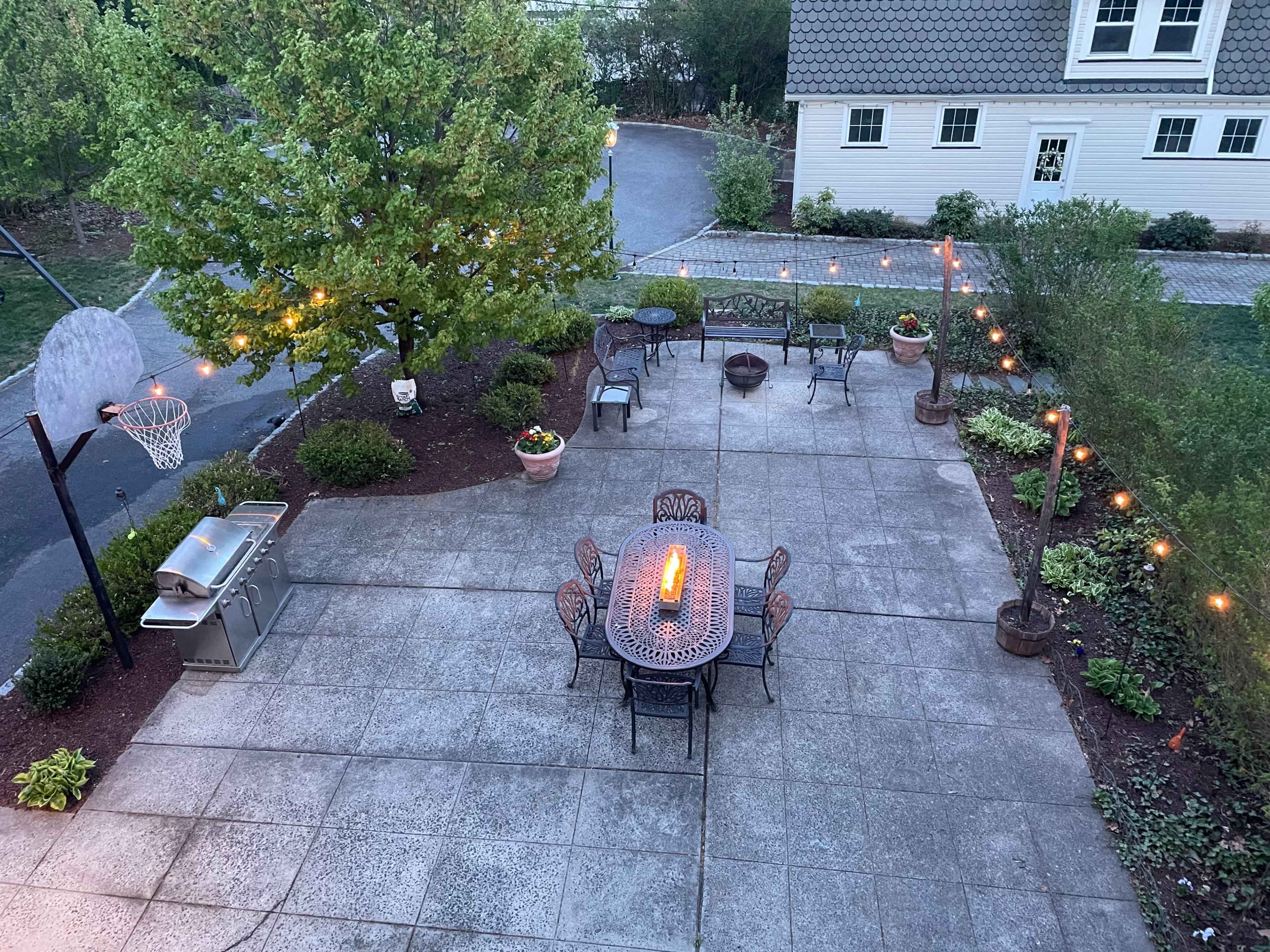 The image shows a spacious backyard patio featuring a basketball hoop, outdoor dining table with chairs, a grill, and garden lights illuminating the area.