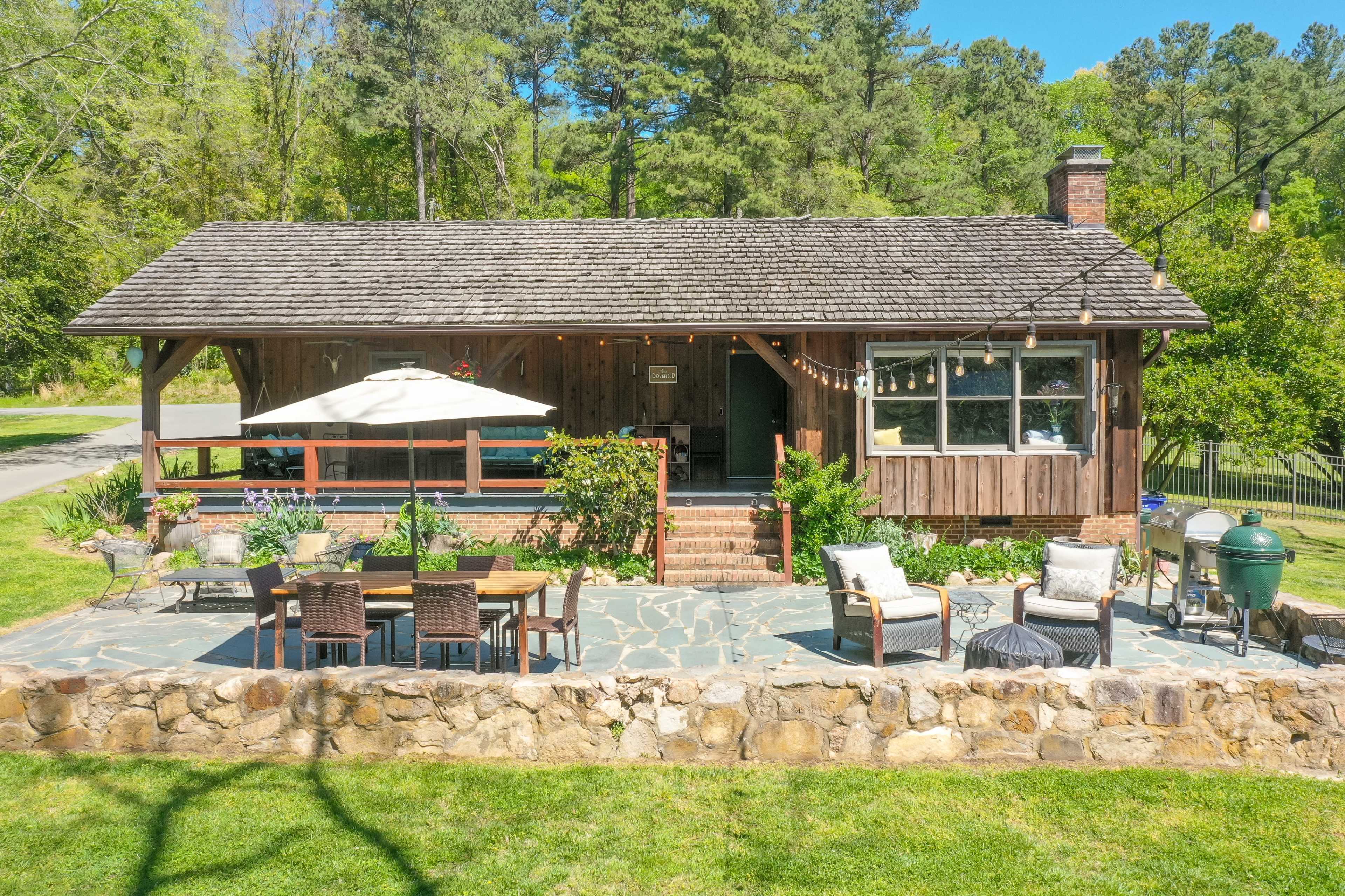 The image shows a cozy wooden cabin with a stone patio, outdoor seating area, and an umbrella, surrounded by greenery.