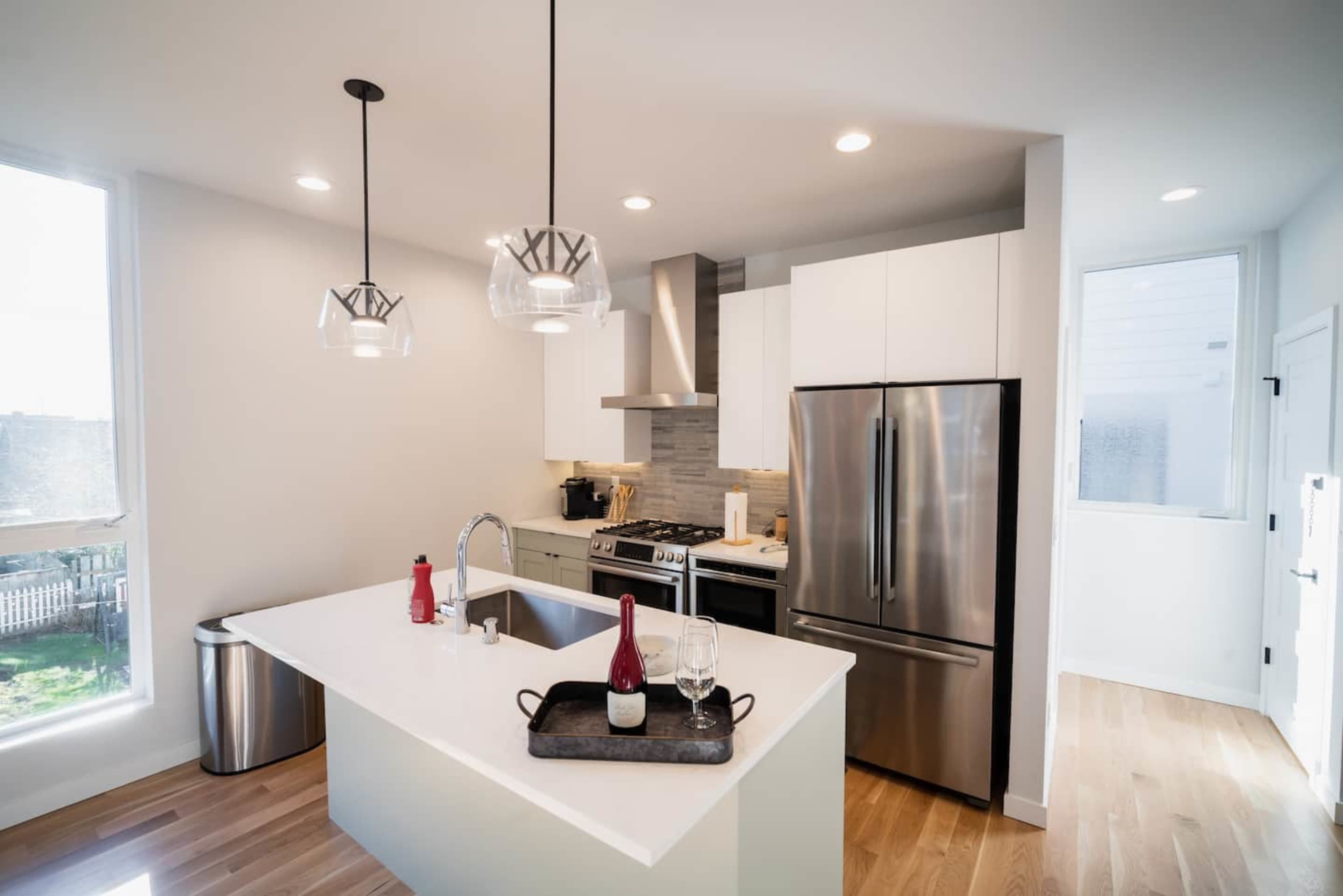 A modern kitchen features a central island with a serving tray, stainless steel appliances, and pendant lighting.