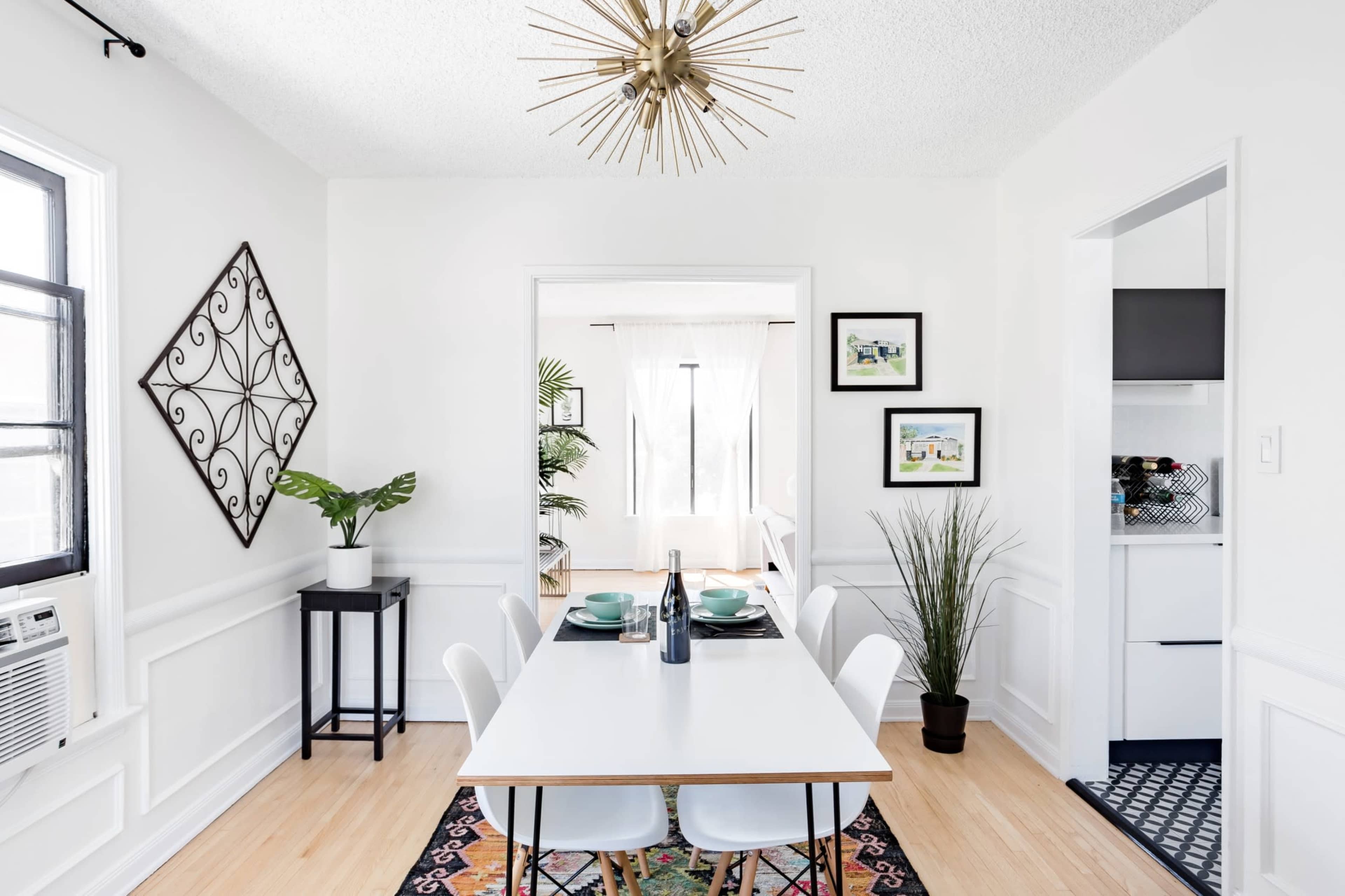 A modern dining area features a white table set for four, surrounded by sleek chairs and accented by decorative plants and wall art.