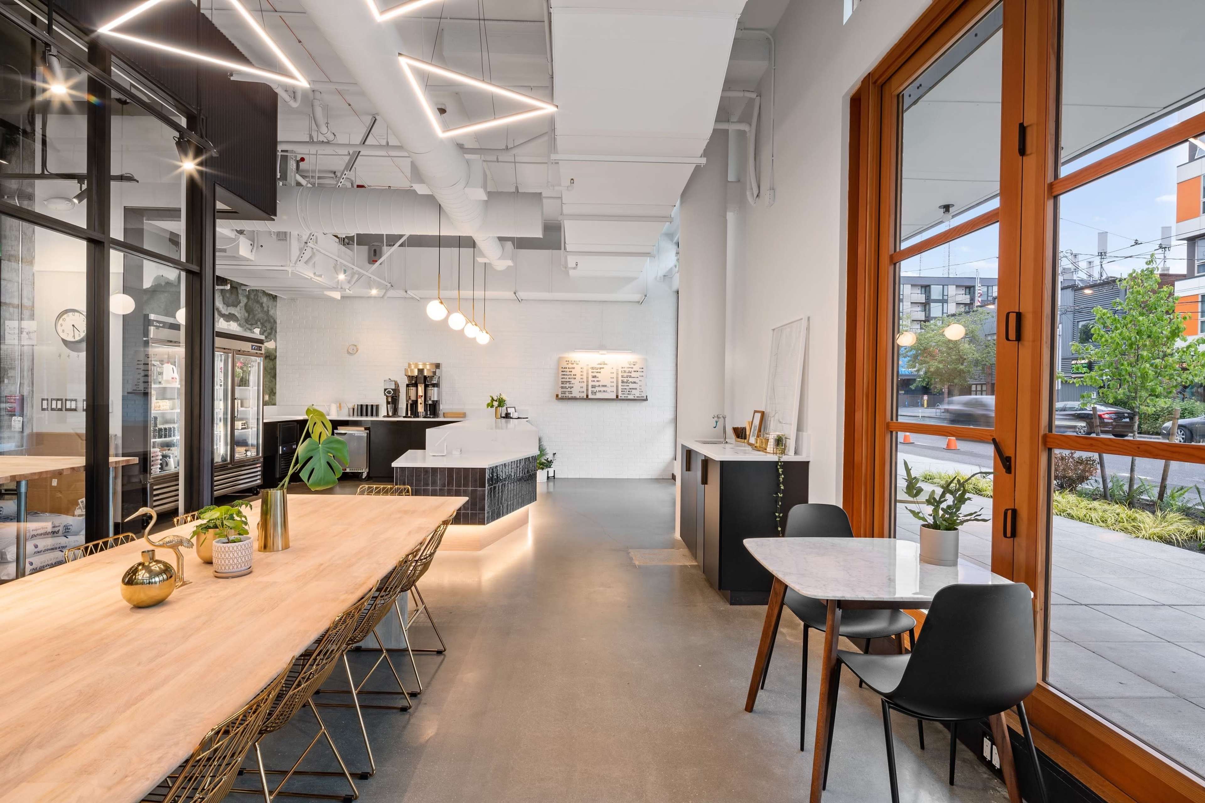 A modern café interior featuring a long wooden table, stylish chairs, and large windows that provide natural light.
