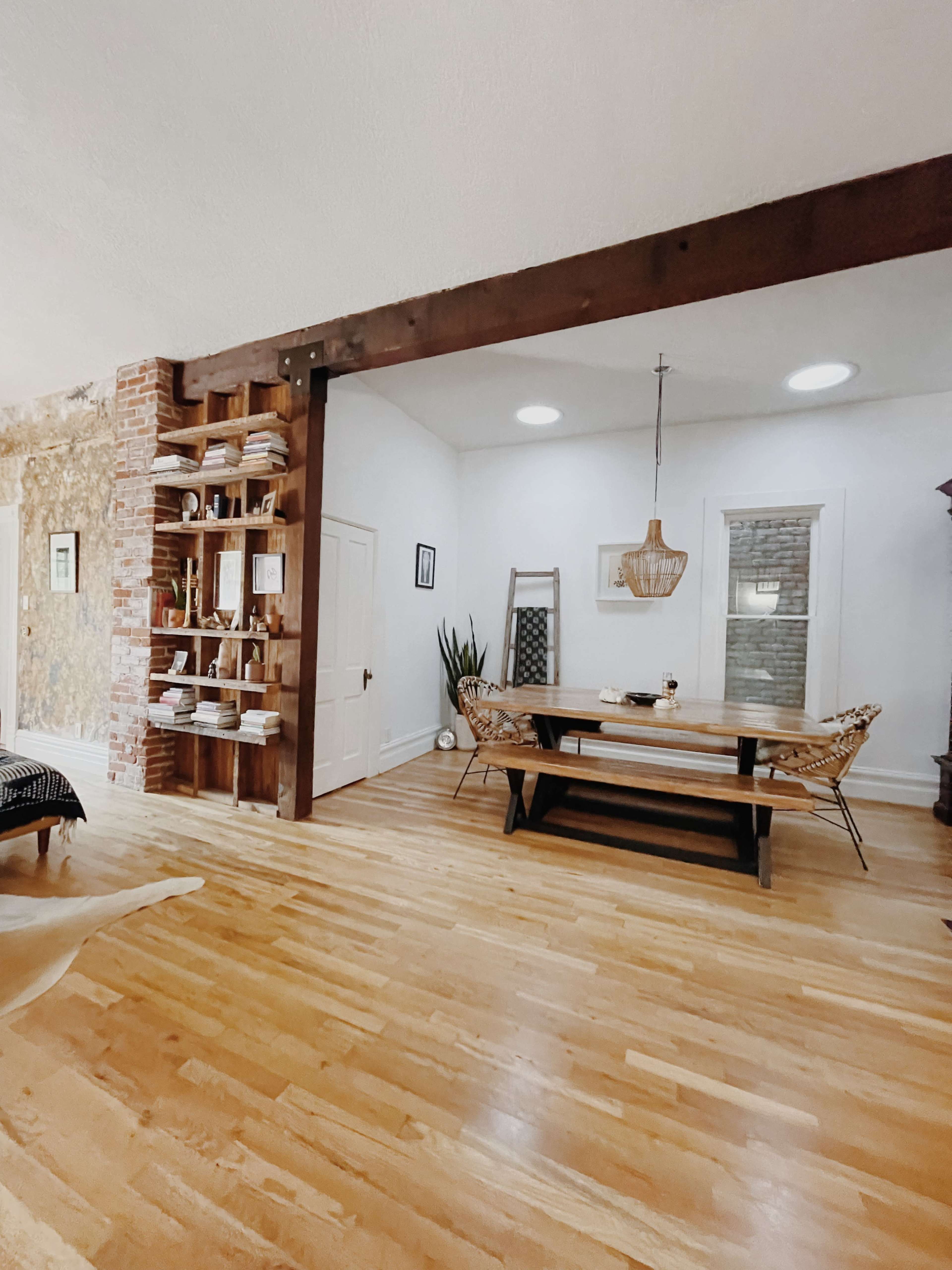 A wooden dining table and chairs are positioned in a bright, open room featuring exposed brick and wooden beams.