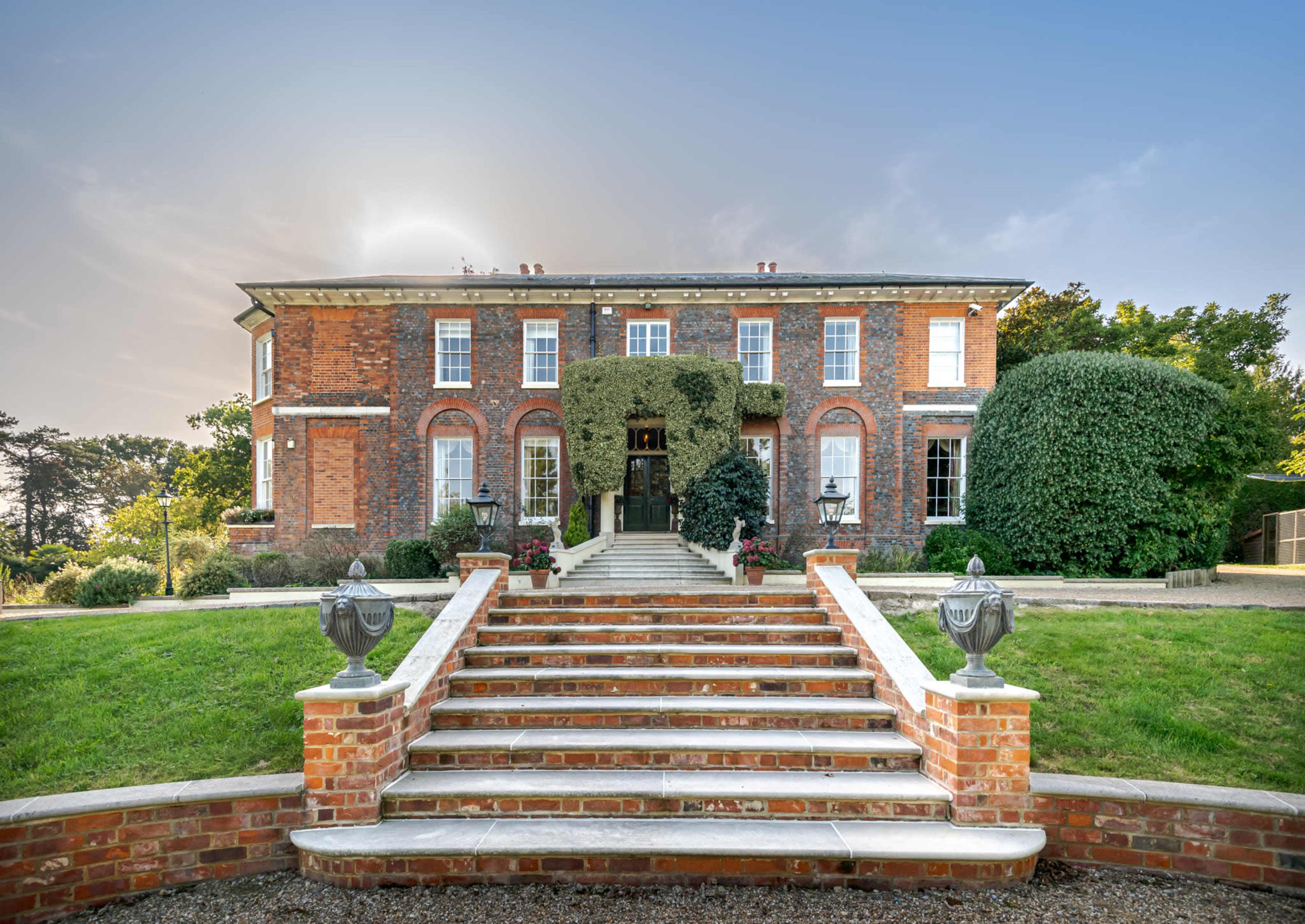A grand brick house with a large staircase and symmetrical landscaping is set against a clear sky.