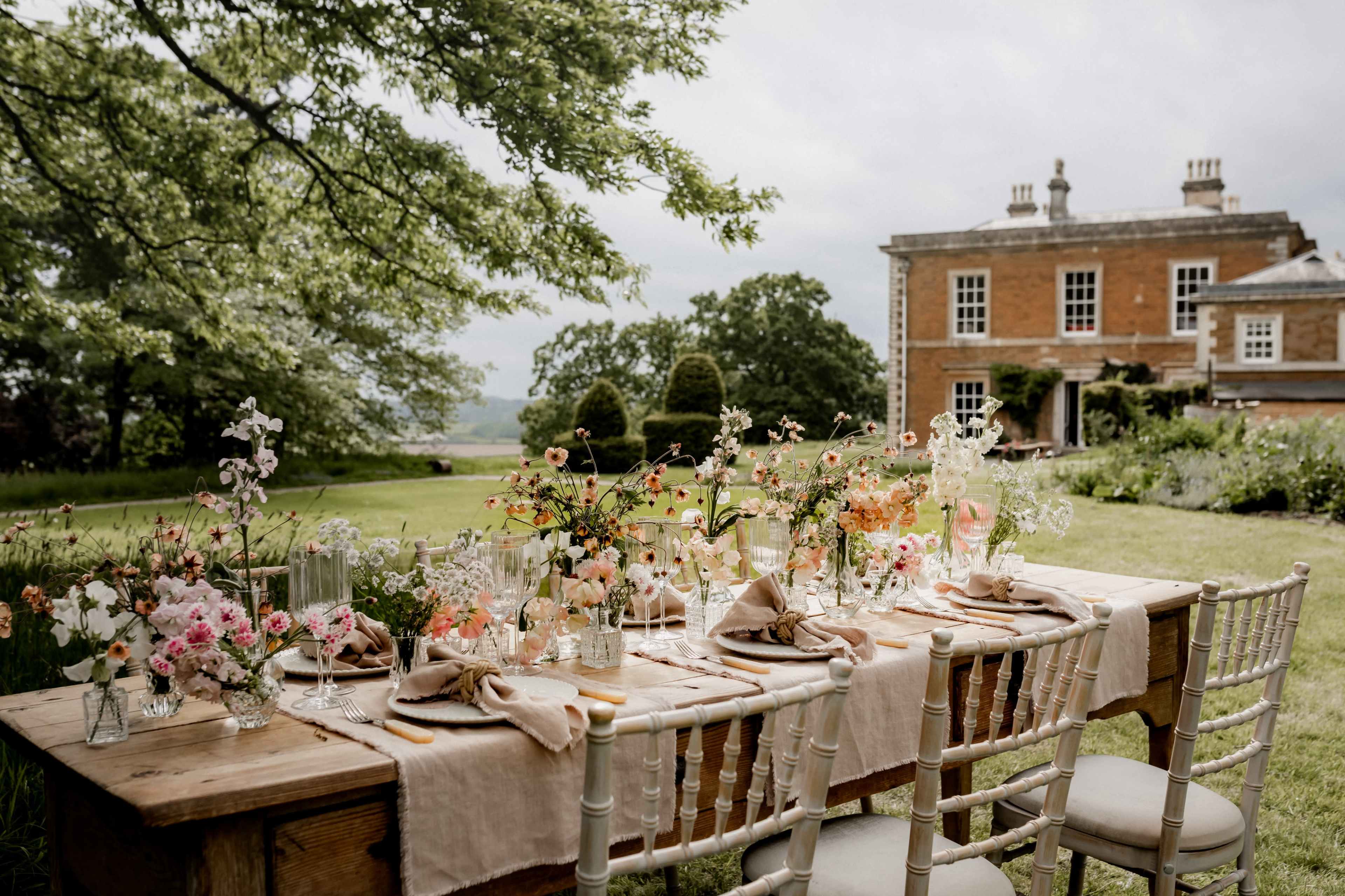 A long wooden table is set outdoors with a variety of flowers, glassware, and linen in front of a large, historic building and lush greenery.