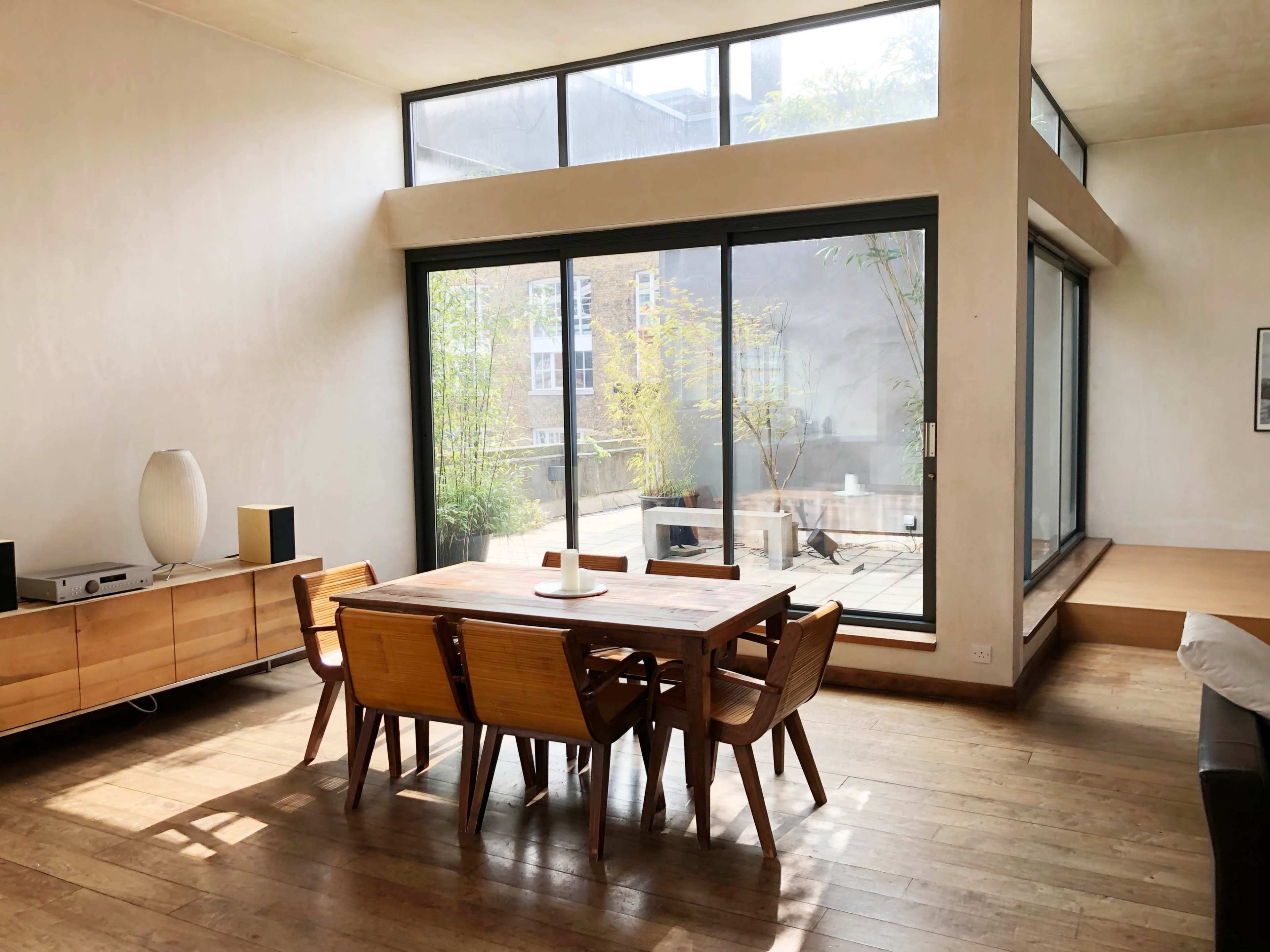 A modern dining area features a wooden table surrounded by chairs, with large glass windows providing a view of an outdoor space filled with greenery.