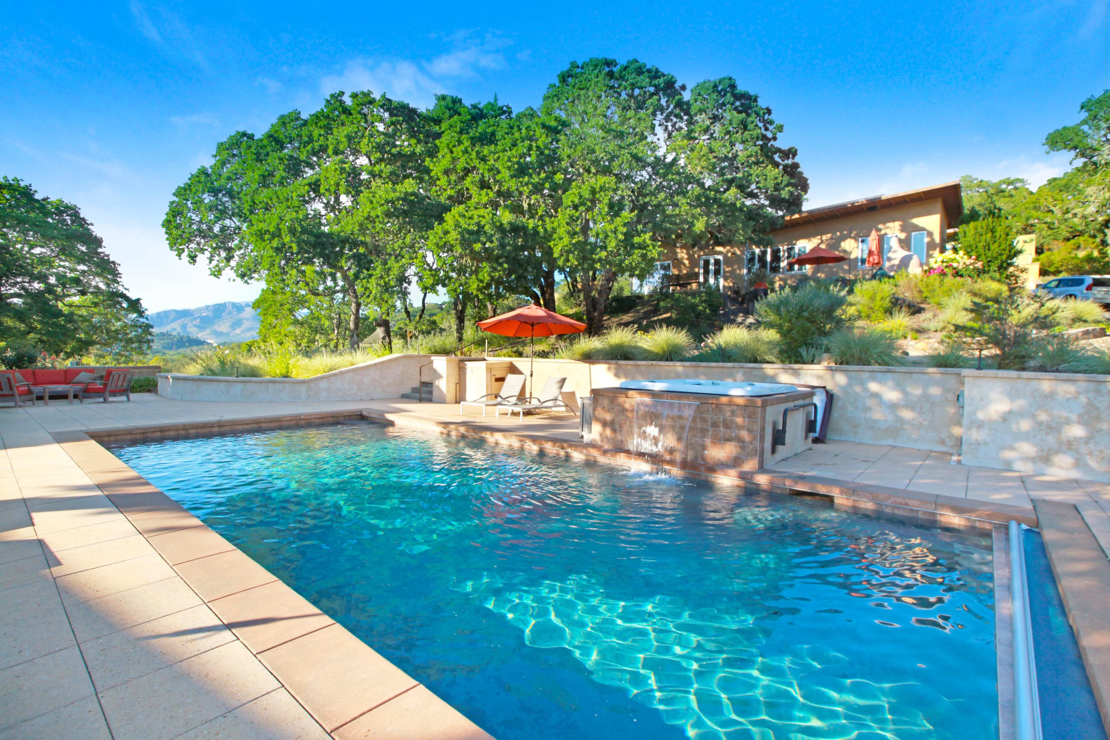 A swimming pool surrounded by a stone patio, with lounge chairs and an umbrella, set against a backdrop of trees and a hillside house.