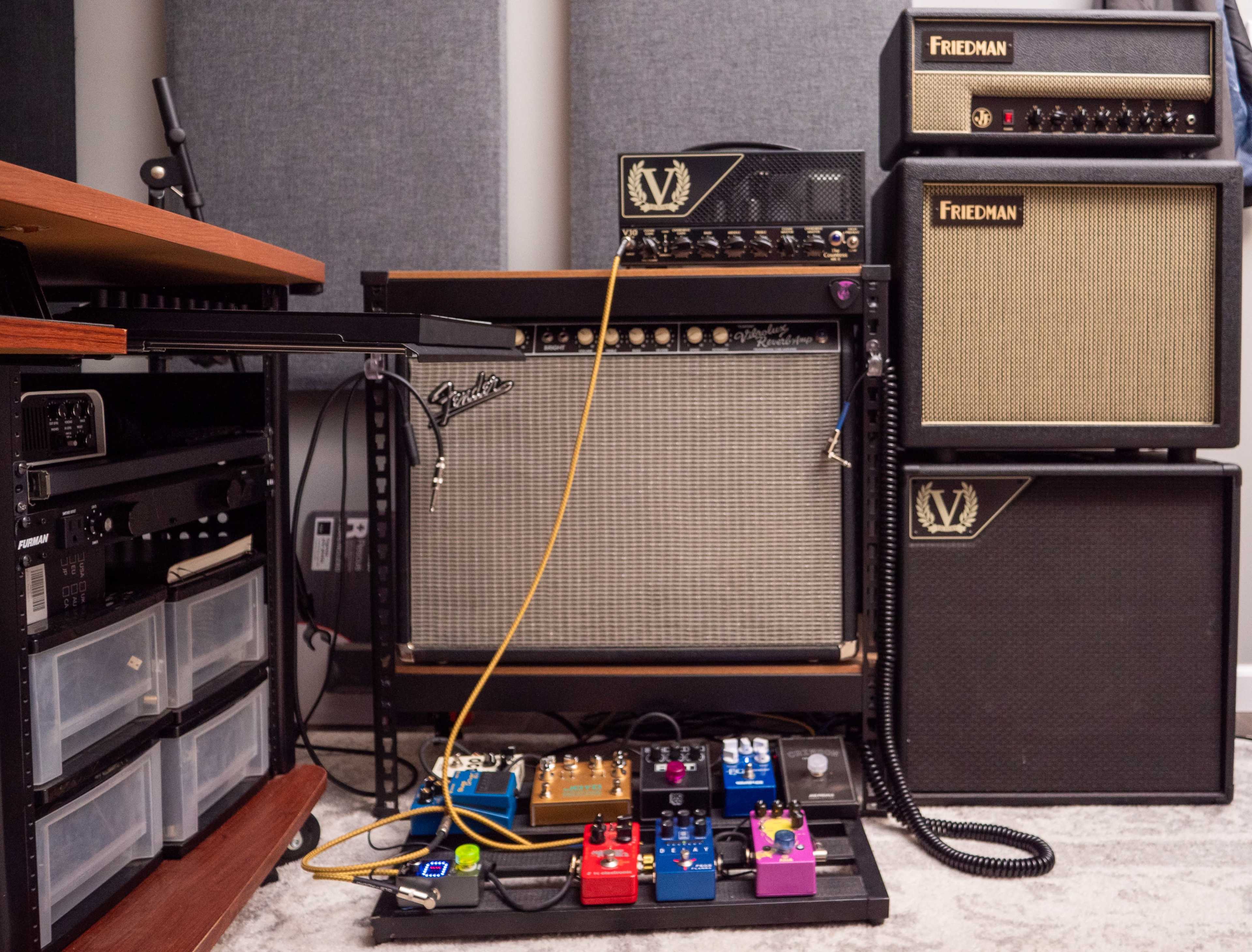 A collection of guitar amplifiers and effects pedals is arranged on a carpeted floor next to a wooden desk in a music studio.