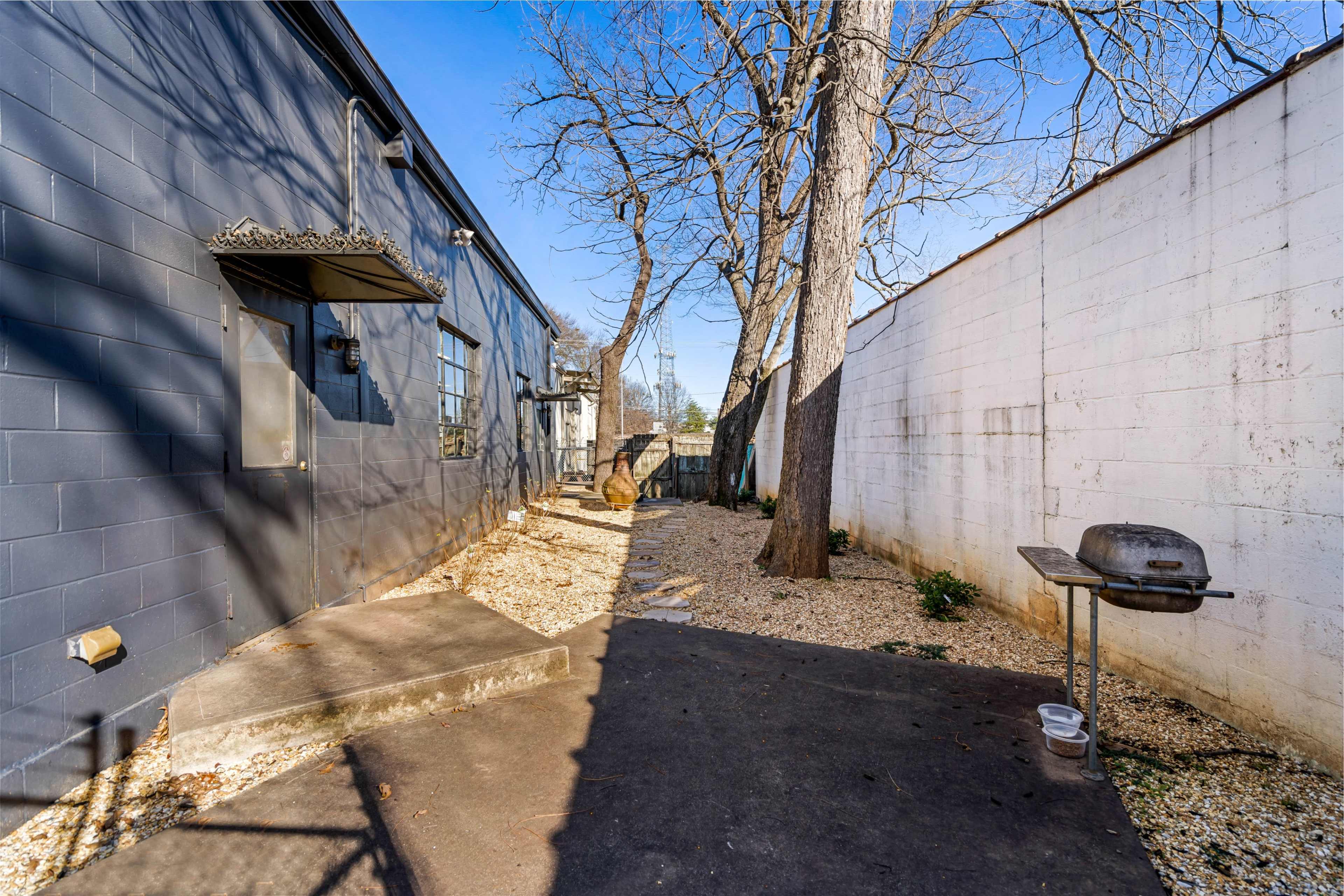 A narrow walkway lined with gravel and trees runs alongside a building, leading to a grill positioned on a concrete slab.
