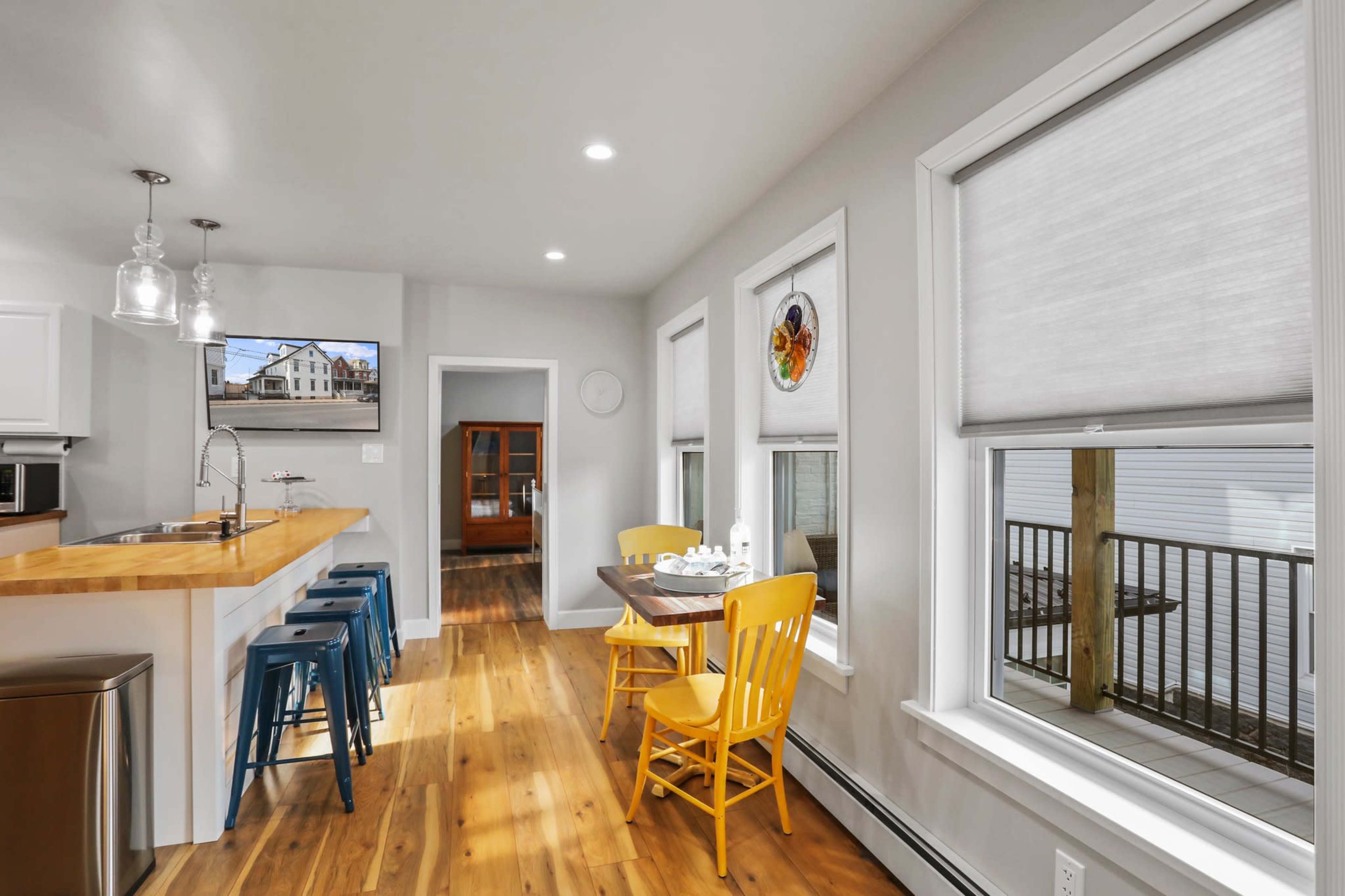 A bright kitchen features a wooden island with bar stools, a dining table with yellow chairs, and large windows overlooking a porch.