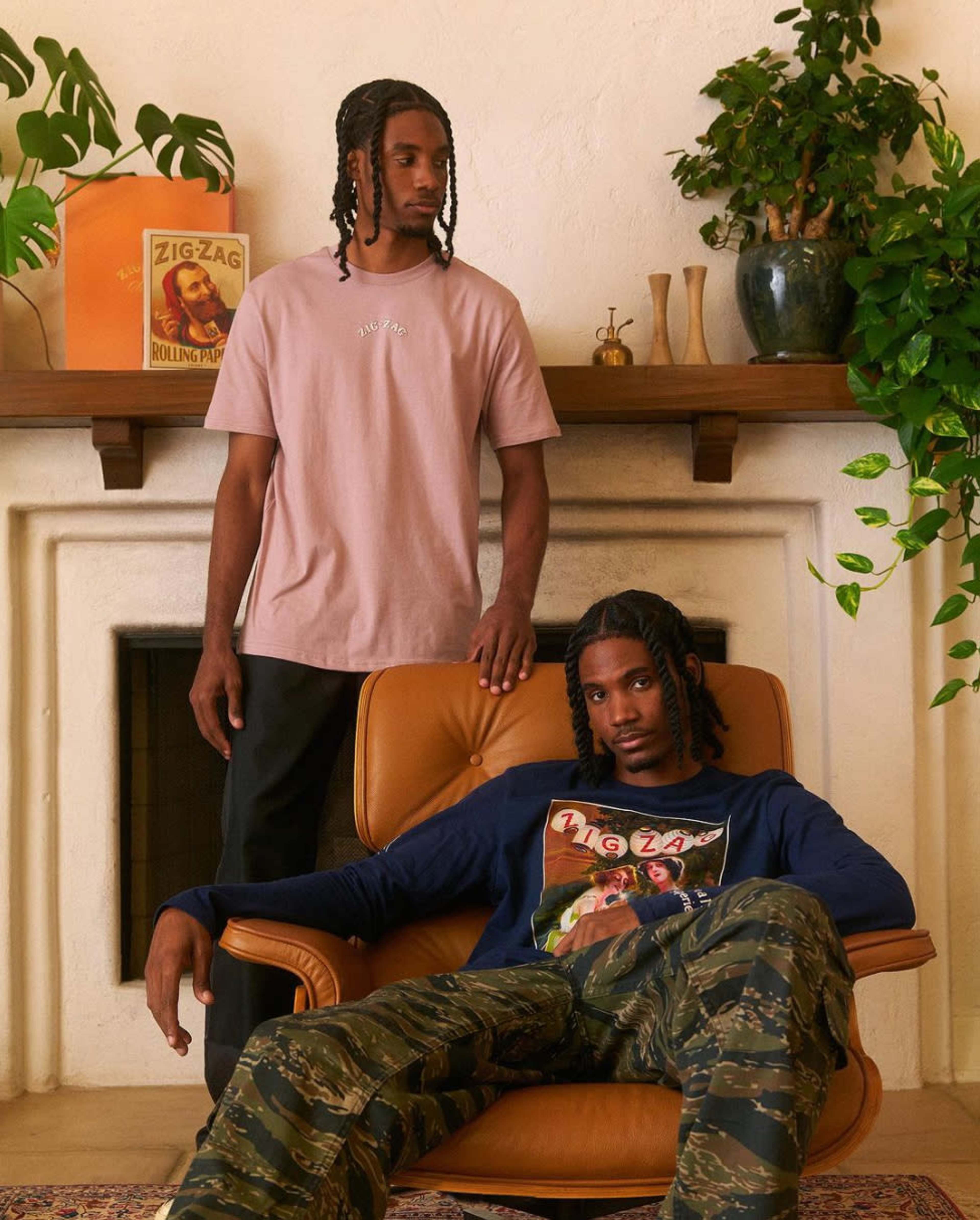 Two men with braided hair pose in a room with a fireplace, one sitting in an orange armchair wearing a graphic sweatshirt and camouflage pants, and the other standing in a pink t-shirt and black pants.