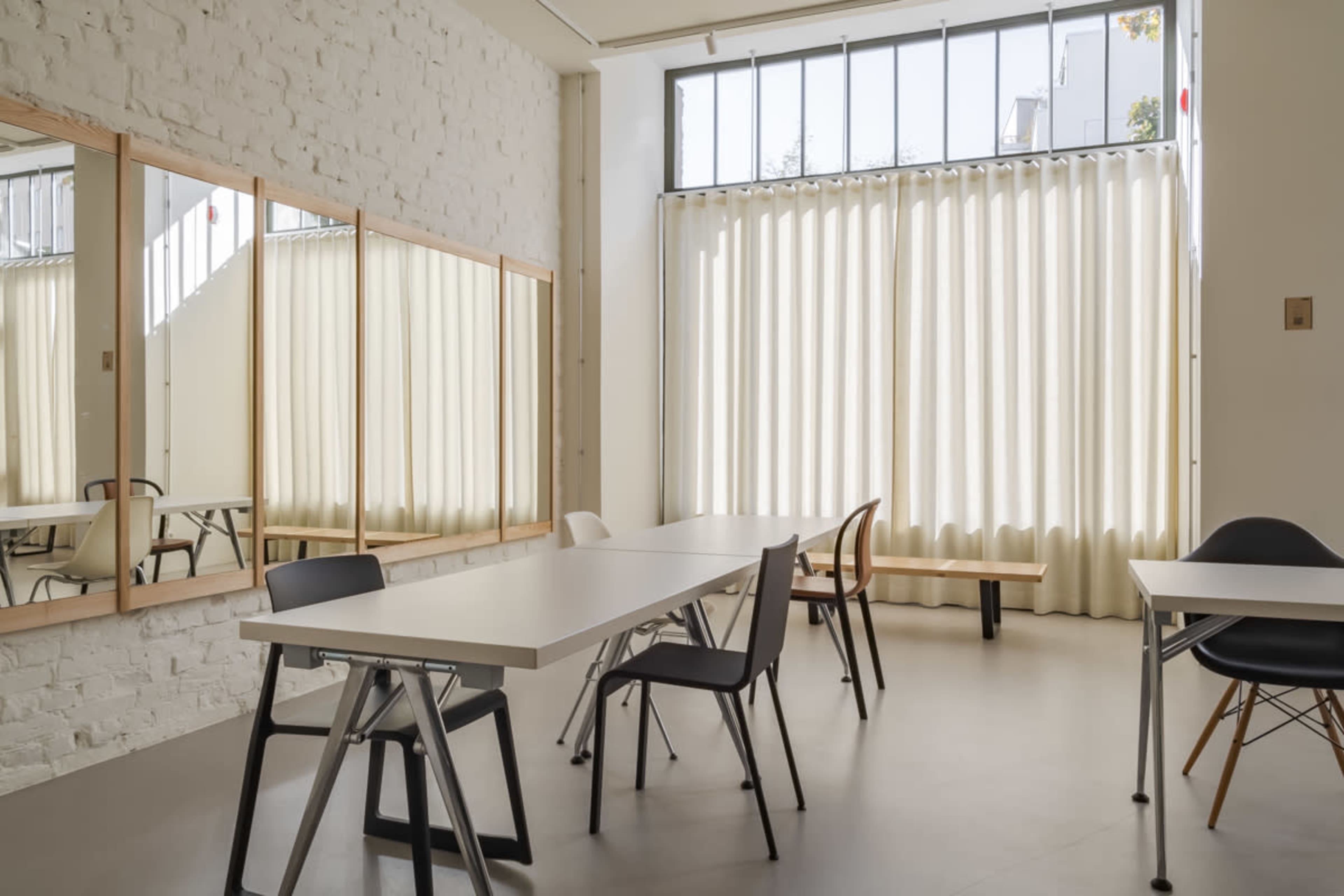 A brightly lit interior space with a large table, several chairs, and two mirrors on the wall, in front of sheer curtains.