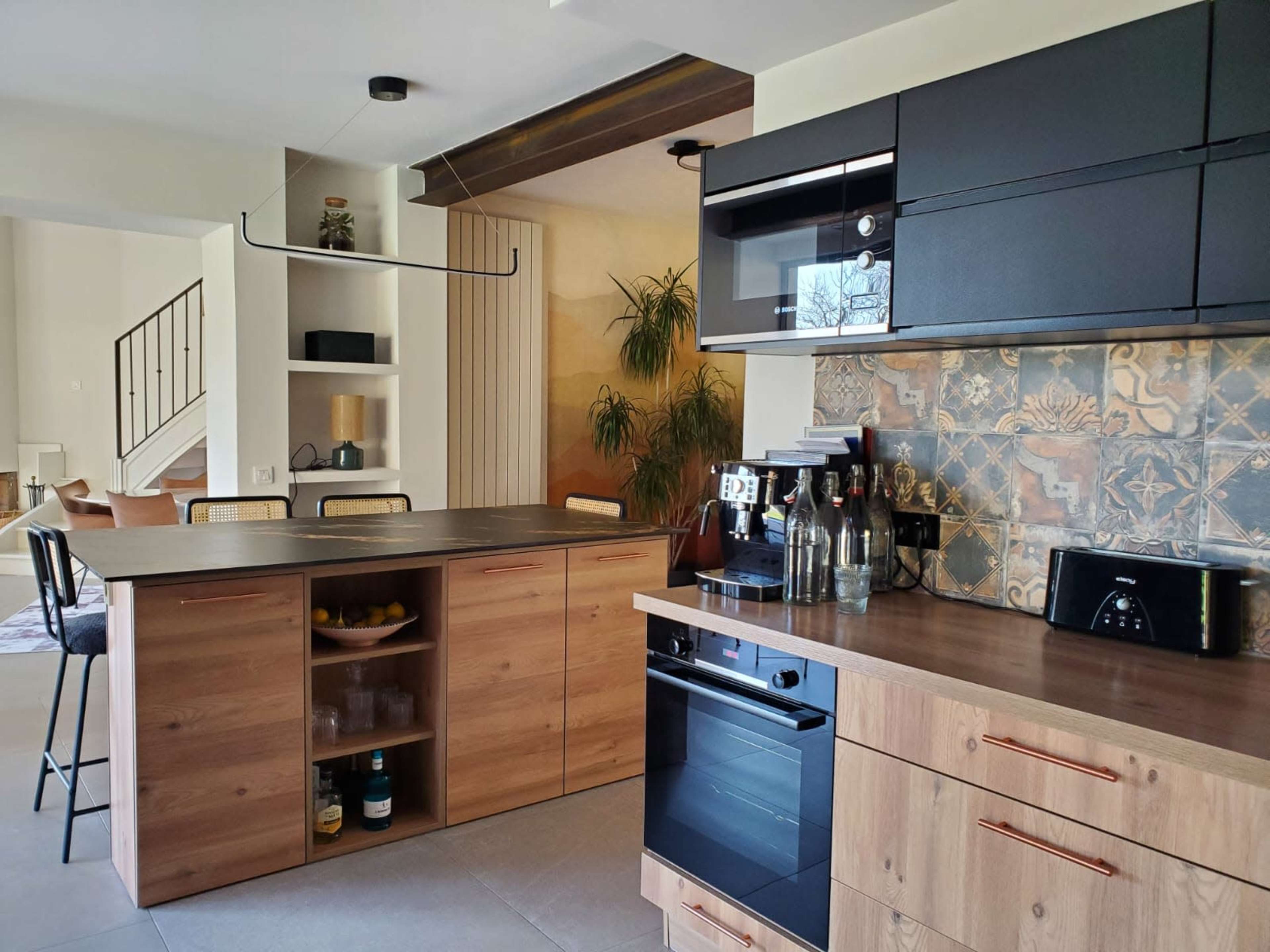 The image shows a modern kitchen with a central island, featuring wood and black cabinets, a decorative tiled backsplash, and various appliances arranged neatly.