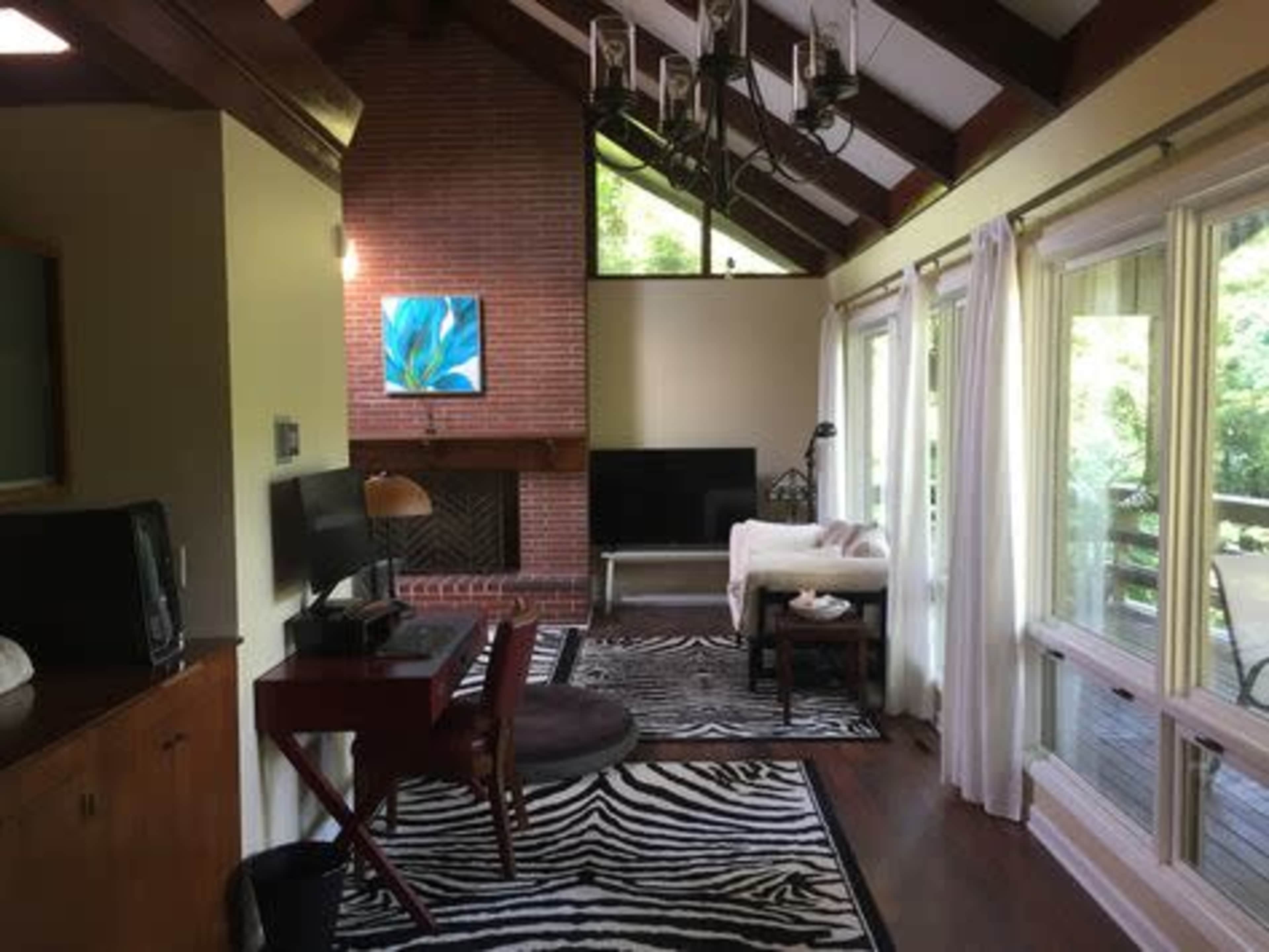 The image shows a living room with a high ceiling, featuring a brick fireplace, a mounted artwork, a television, and large windows leading to a wooden deck.