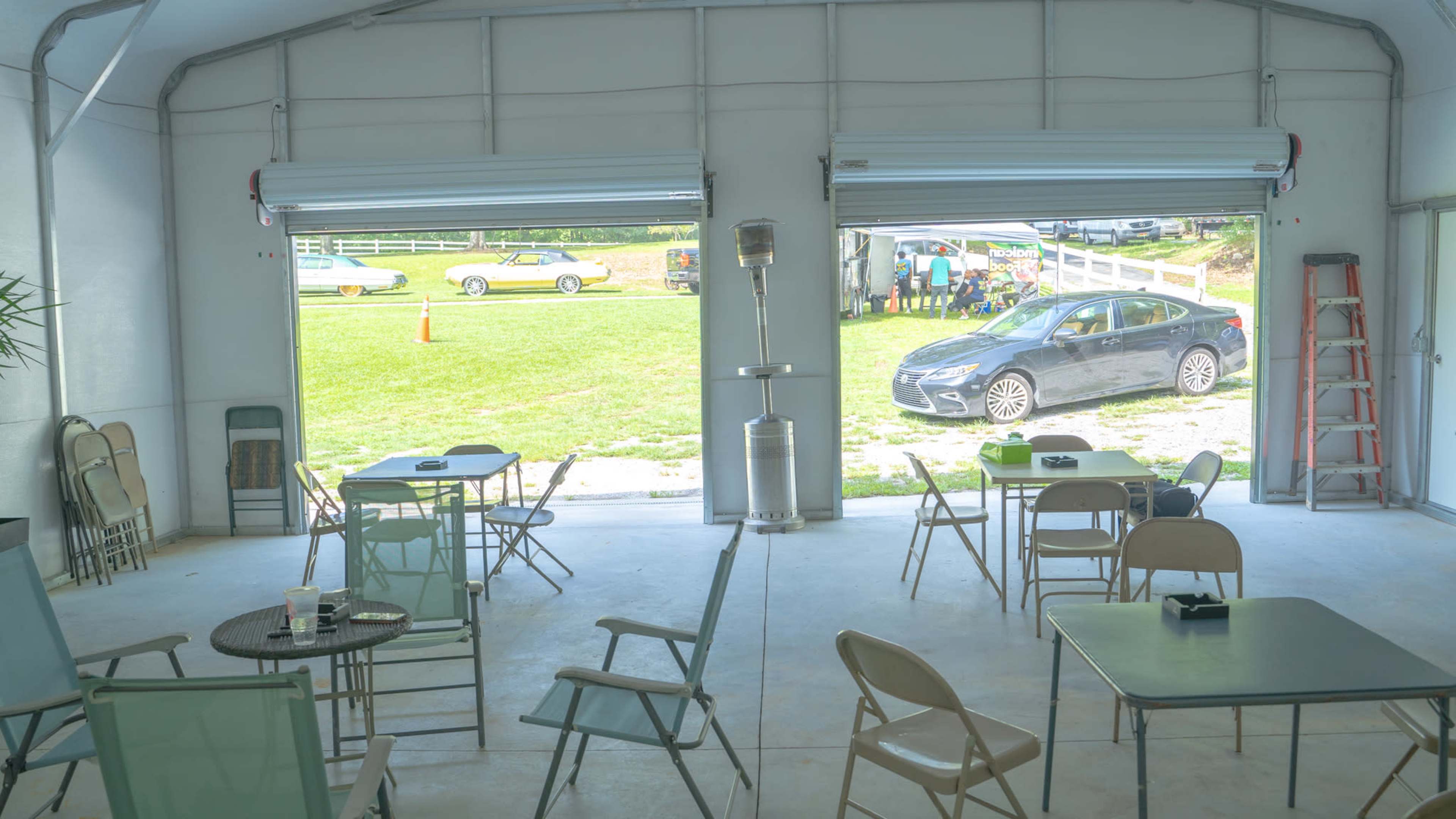 An open garage-like space with tables and chairs arranged inside, while a car is parked outside on the lawn.