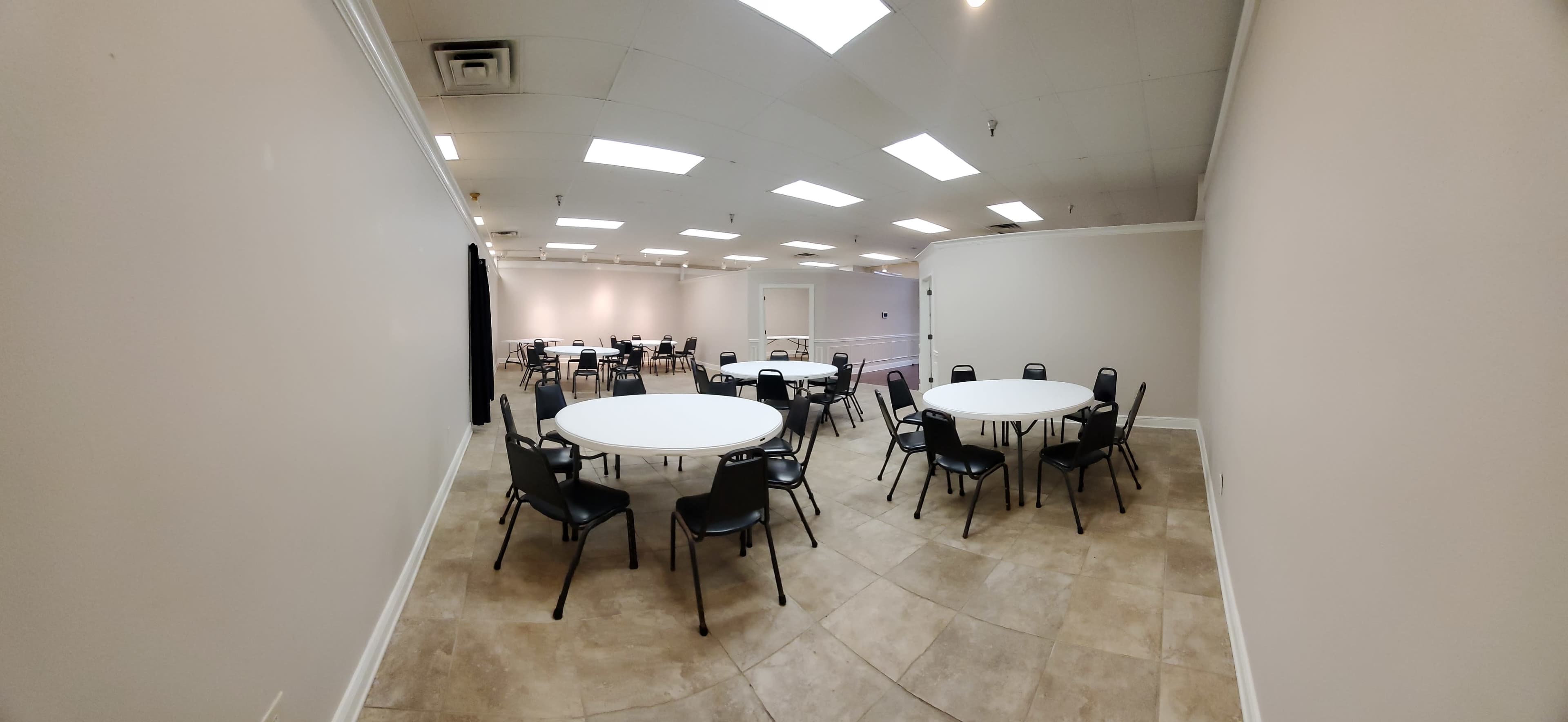 A spacious room with several round tables and chairs arranged across a tiled floor, illuminated by overhead lights.