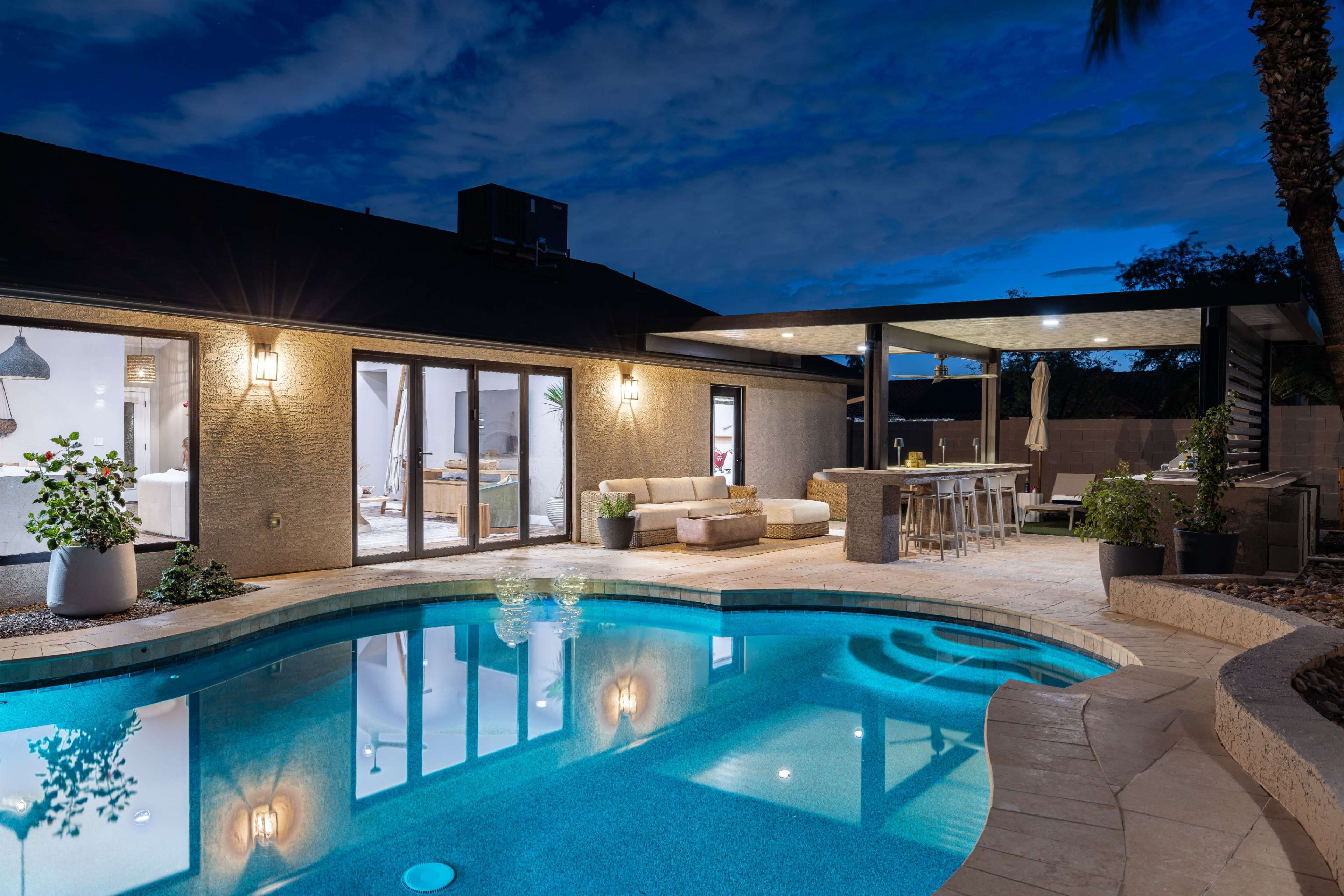The image shows a modern backyard pool area with a seating arrangement and an outdoor dining space, illuminated under a twilight sky.