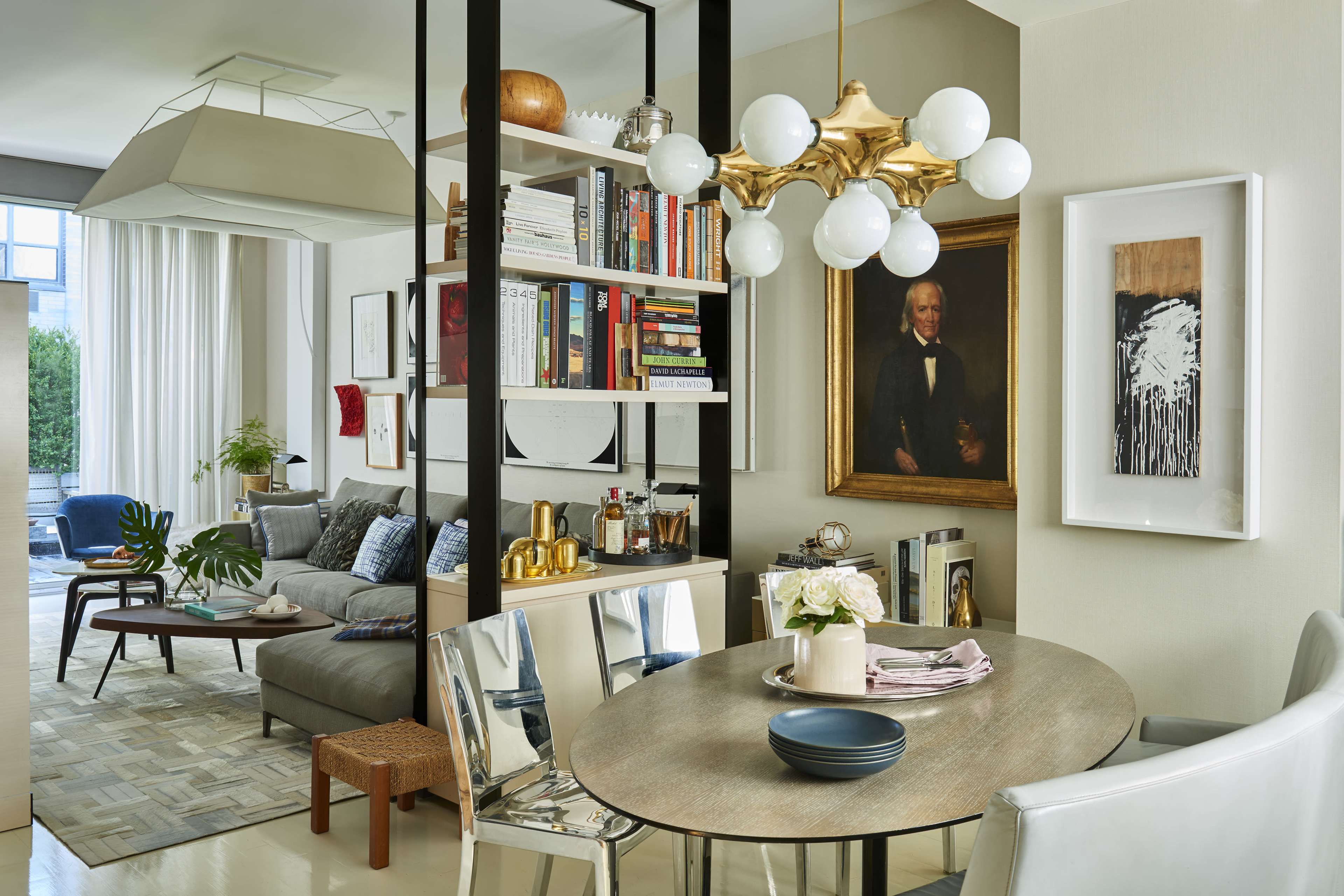 A modern living space with a dining area featuring a round table, a bookshelf, a framed portrait, and a comfortable seating area visible through an open layout.