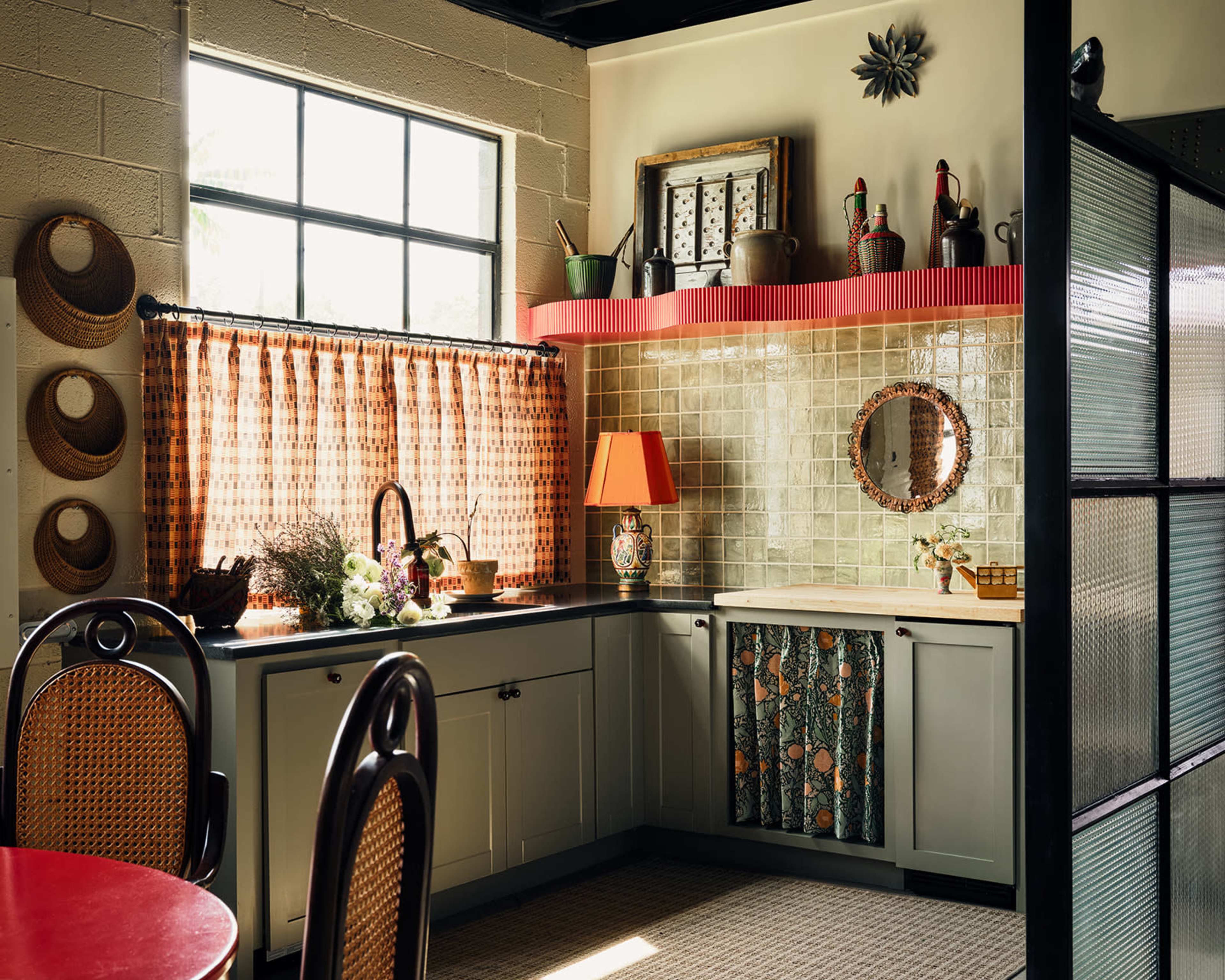 The image shows a kitchen with a window featuring a checkered curtain, a variety of decorative items on the shelves, and a table set with a red surface.