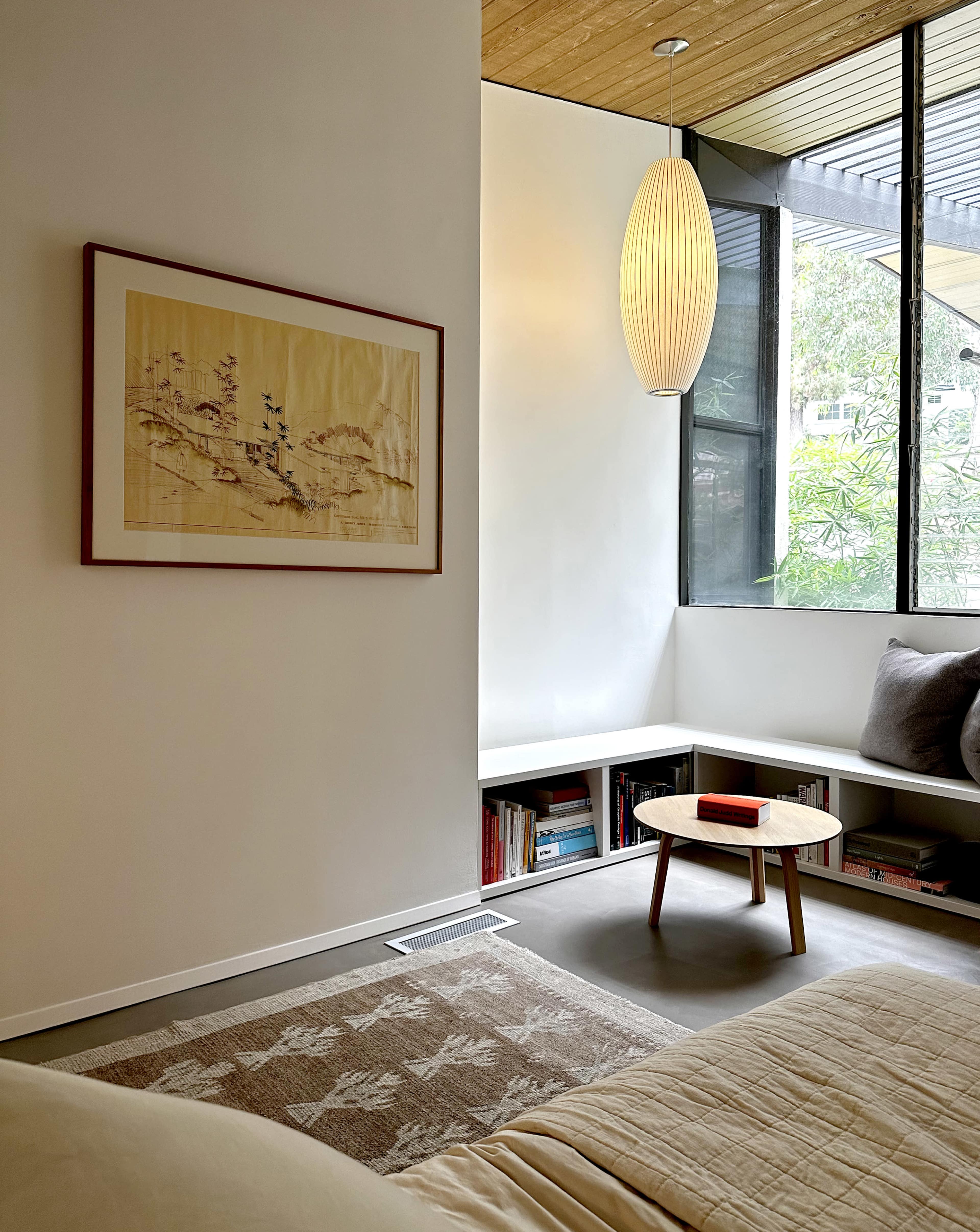 The image shows a minimalist living space featuring a framed artwork, a round coffee table, and a corner window with natural light.