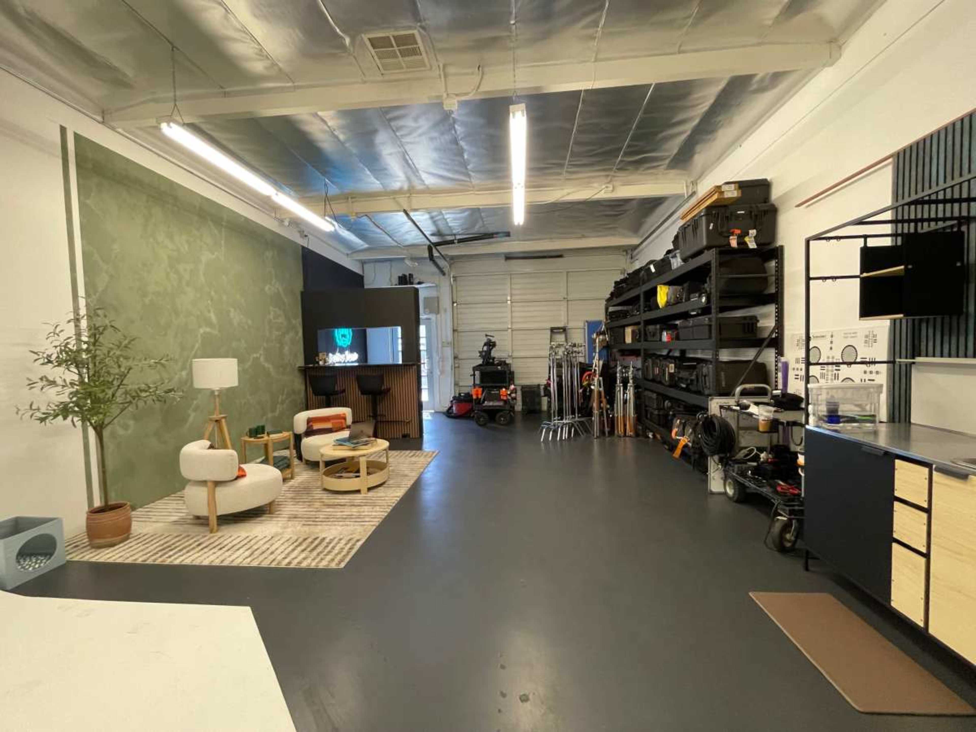 The image shows a spacious studio setting with a marked area featuring modern furniture, including a couch and a coffee table, and shelves stocked with various equipment along one wall.