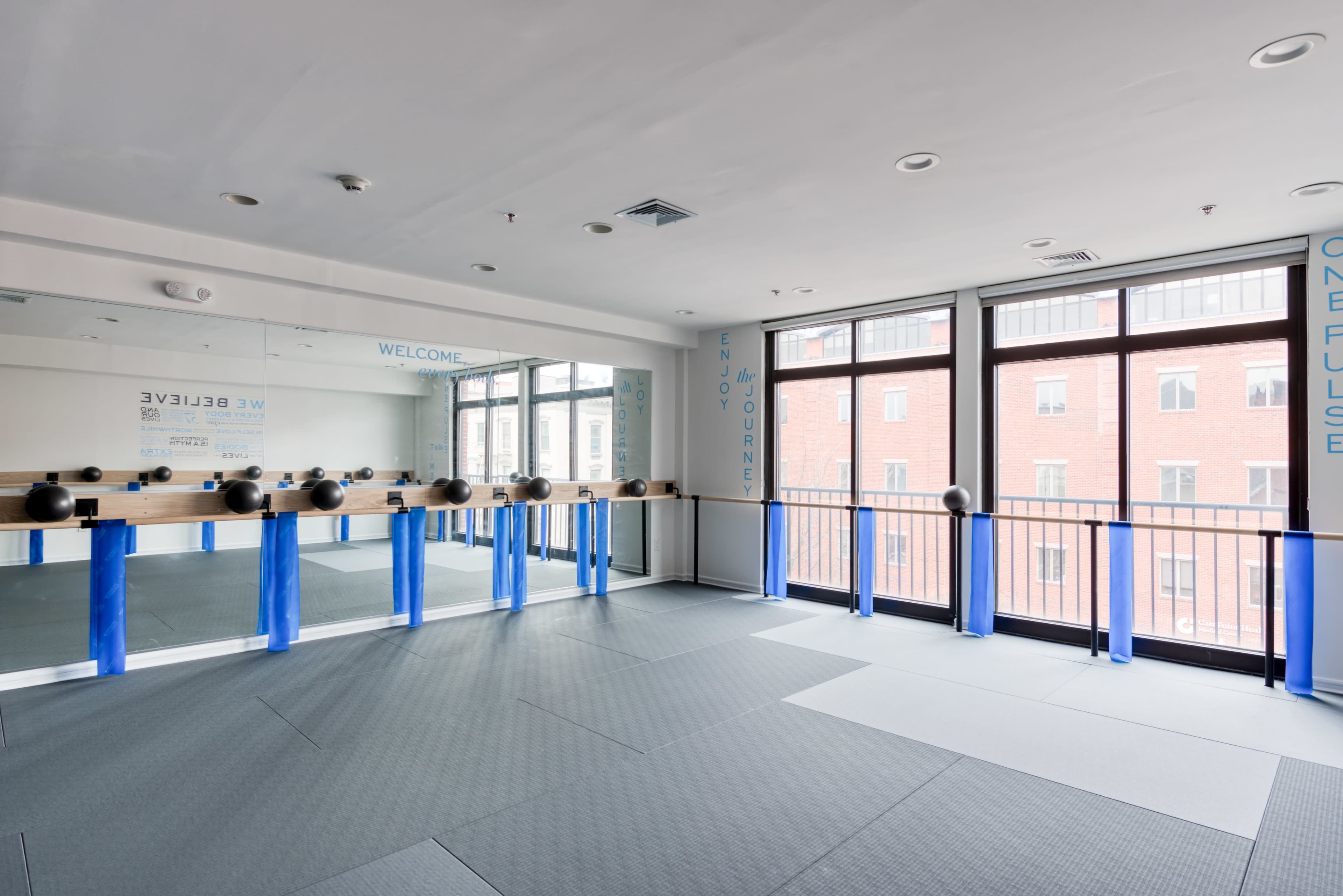 The image shows a bright fitness studio with large windows, mirrored walls, and exercise equipment arranged along a wooden barre.