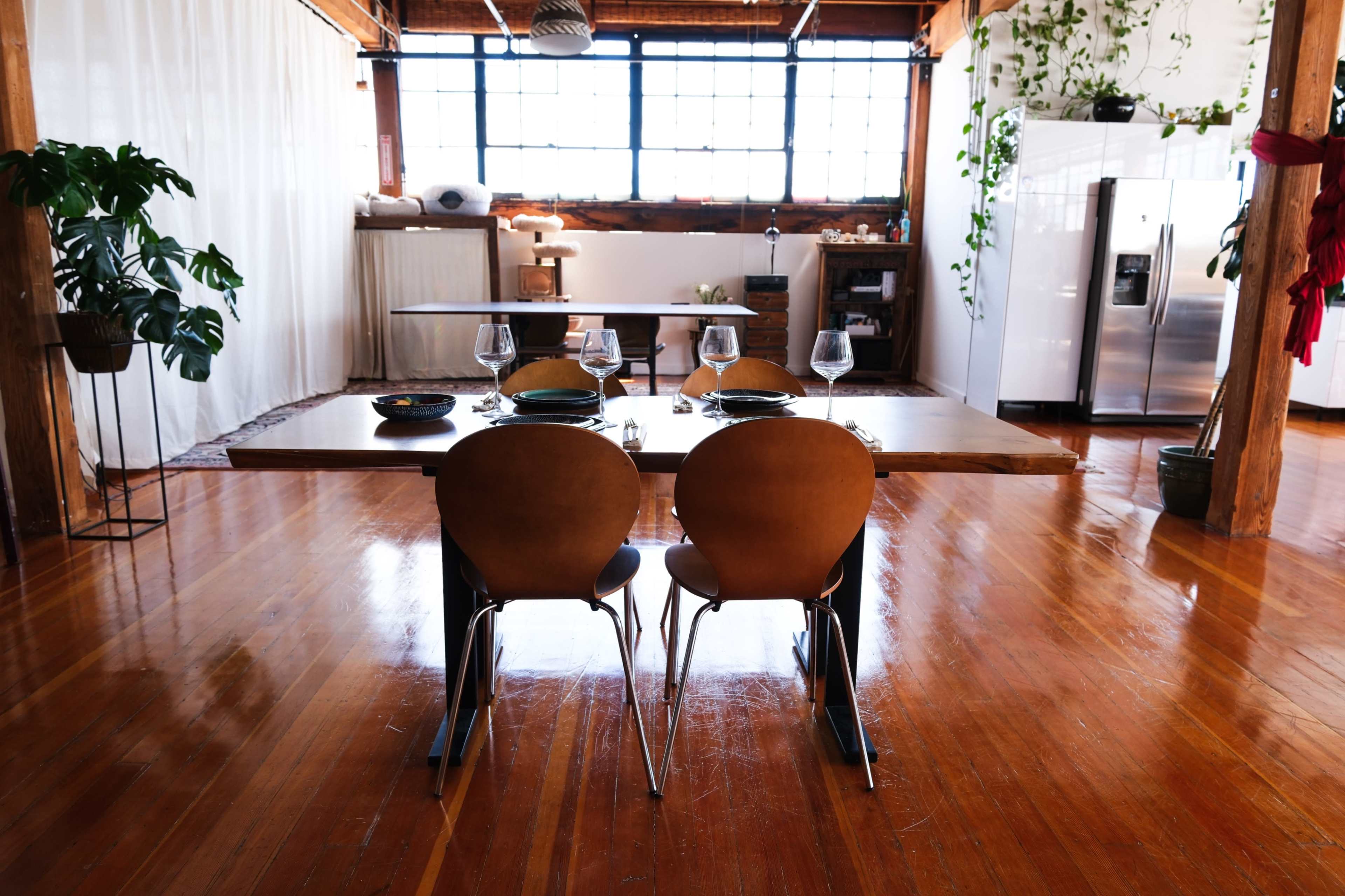 The image shows a spacious dining area with a wooden table set for a meal, complemented by two chairs and large windows letting in natural light.
