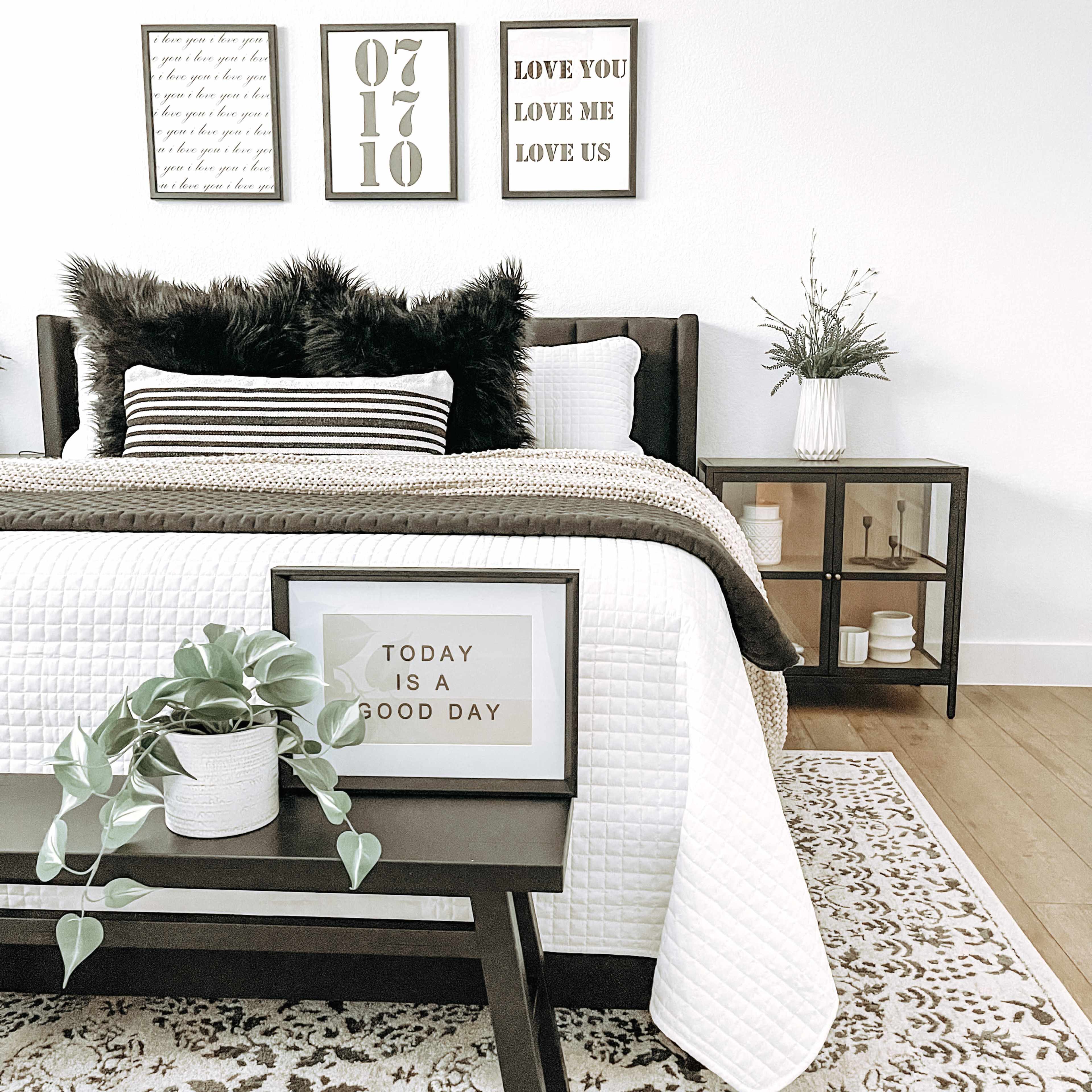 A neatly made bed with decorative pillows is positioned beside a nightstand that holds a framed quote and a small plant, set against a minimalist room with wall art.