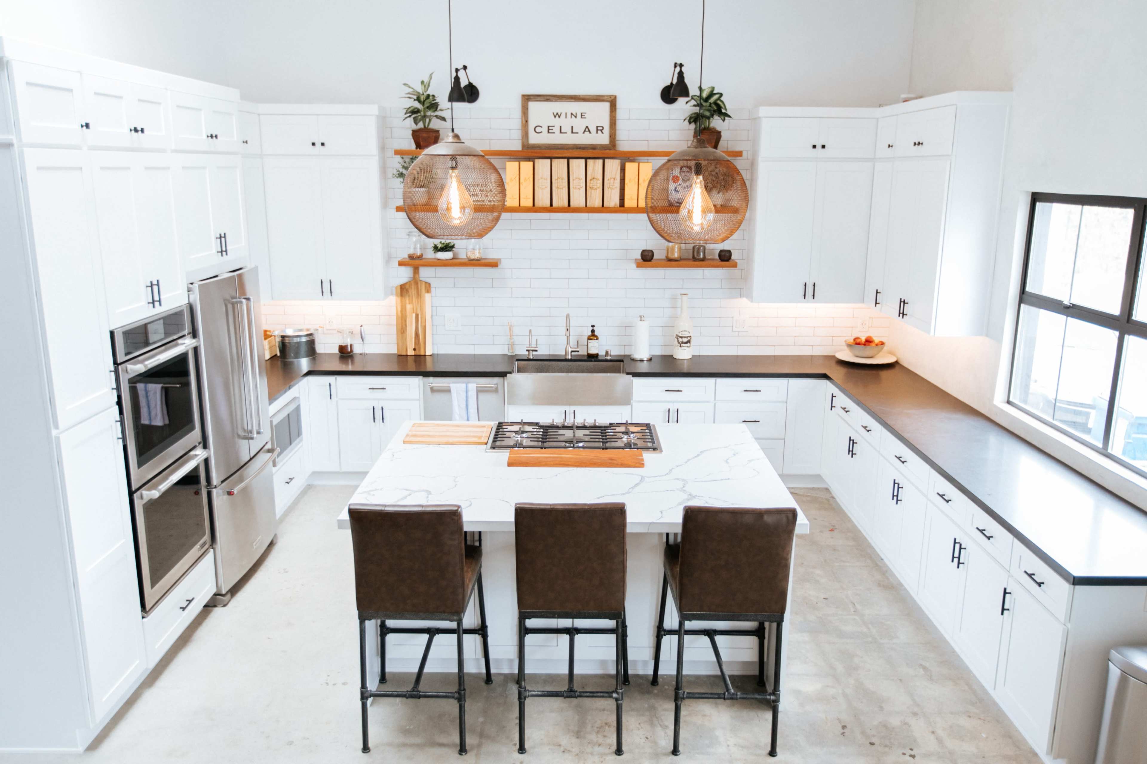 A modern kitchen features a central island with barstools, white cabinetry, a dark countertop, and pendant lighting above, with a wine cellar displayed on the wall.