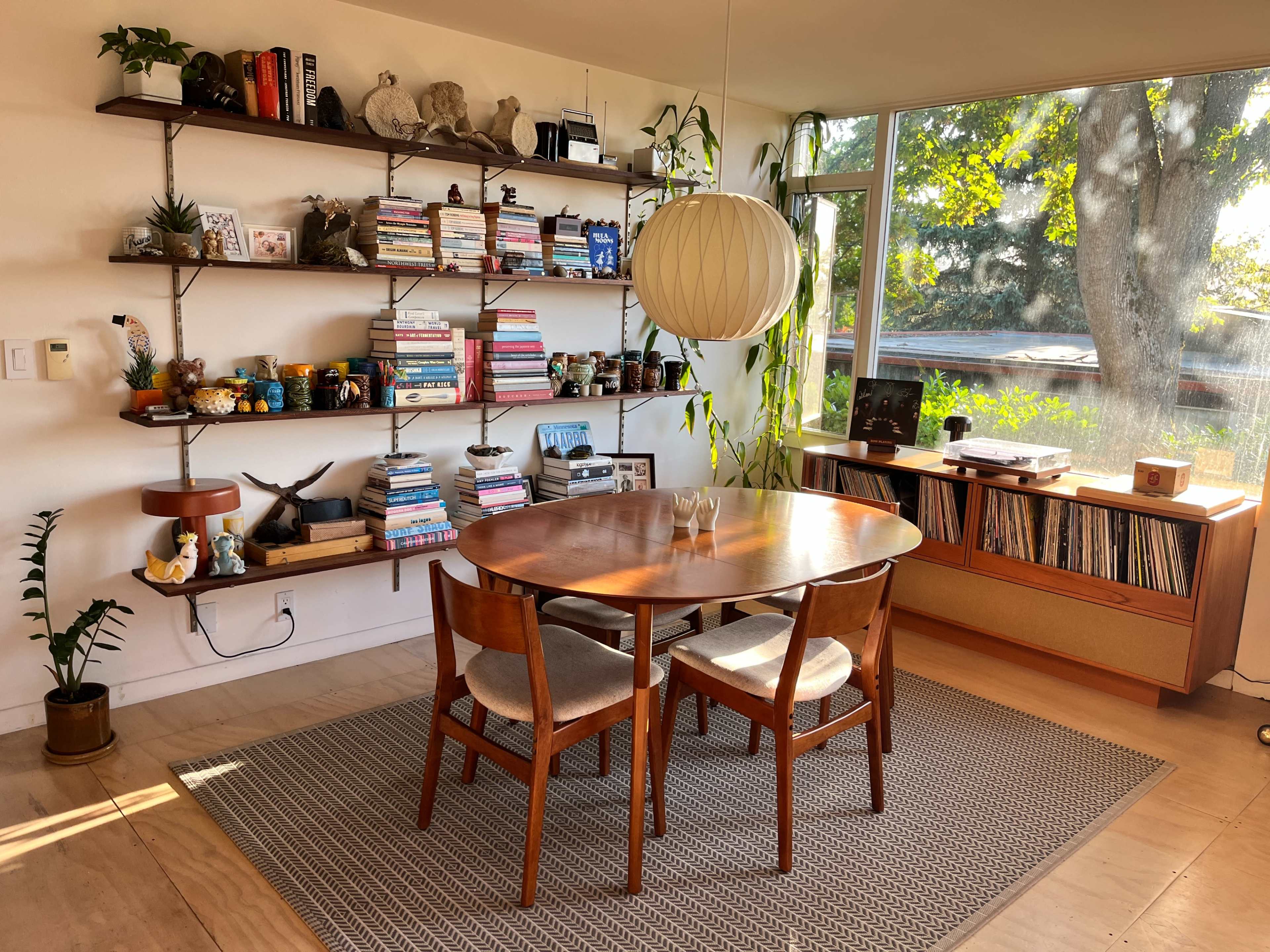A wooden dining table with three chairs is positioned in a well-lit room featuring a large window, surrounded by shelves filled with books and decorative items.