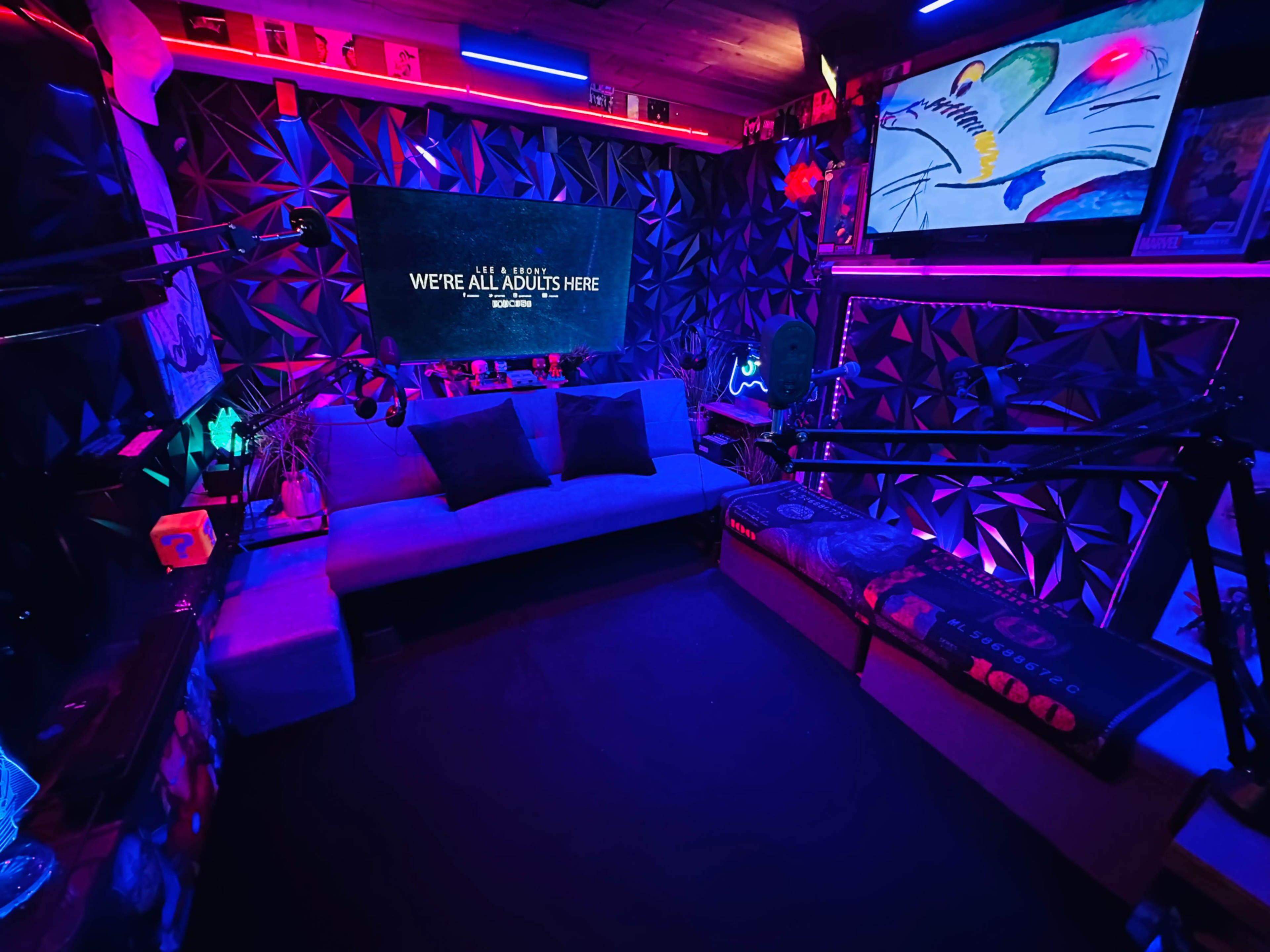 The image shows a dimly lit room with colorful LED lights, featuring a blue couch, a large screen displaying text, and various decorative items on the walls.