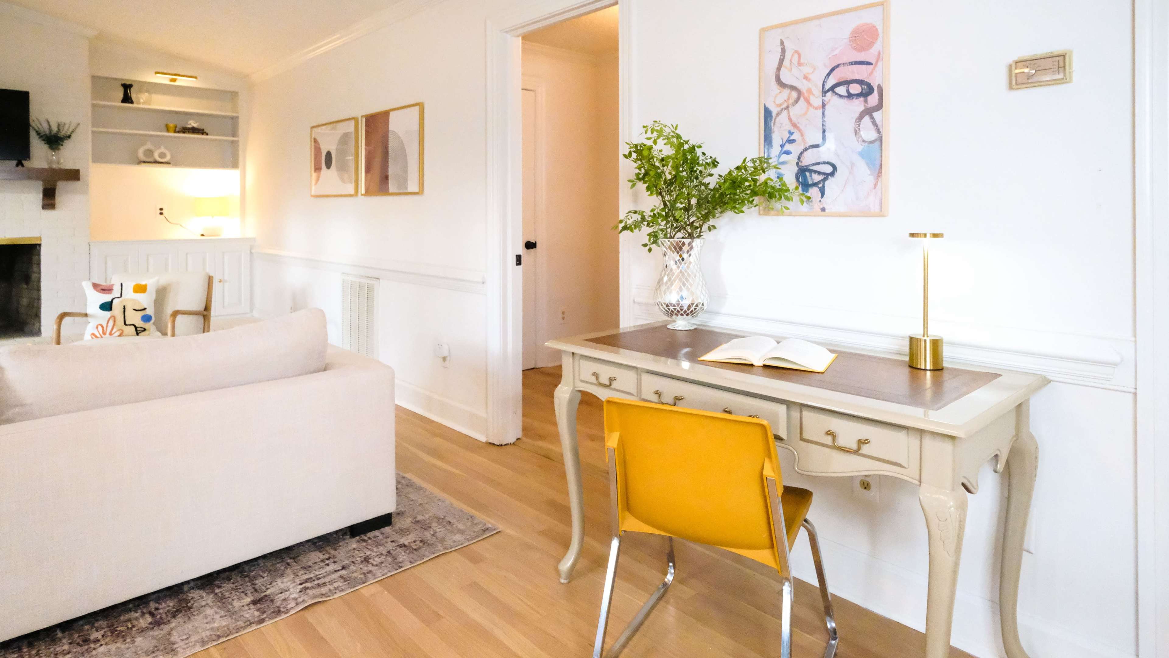 A cozy living area with a beige sofa, a small writing desk paired with a yellow chair, and artwork hanging on the white walls.