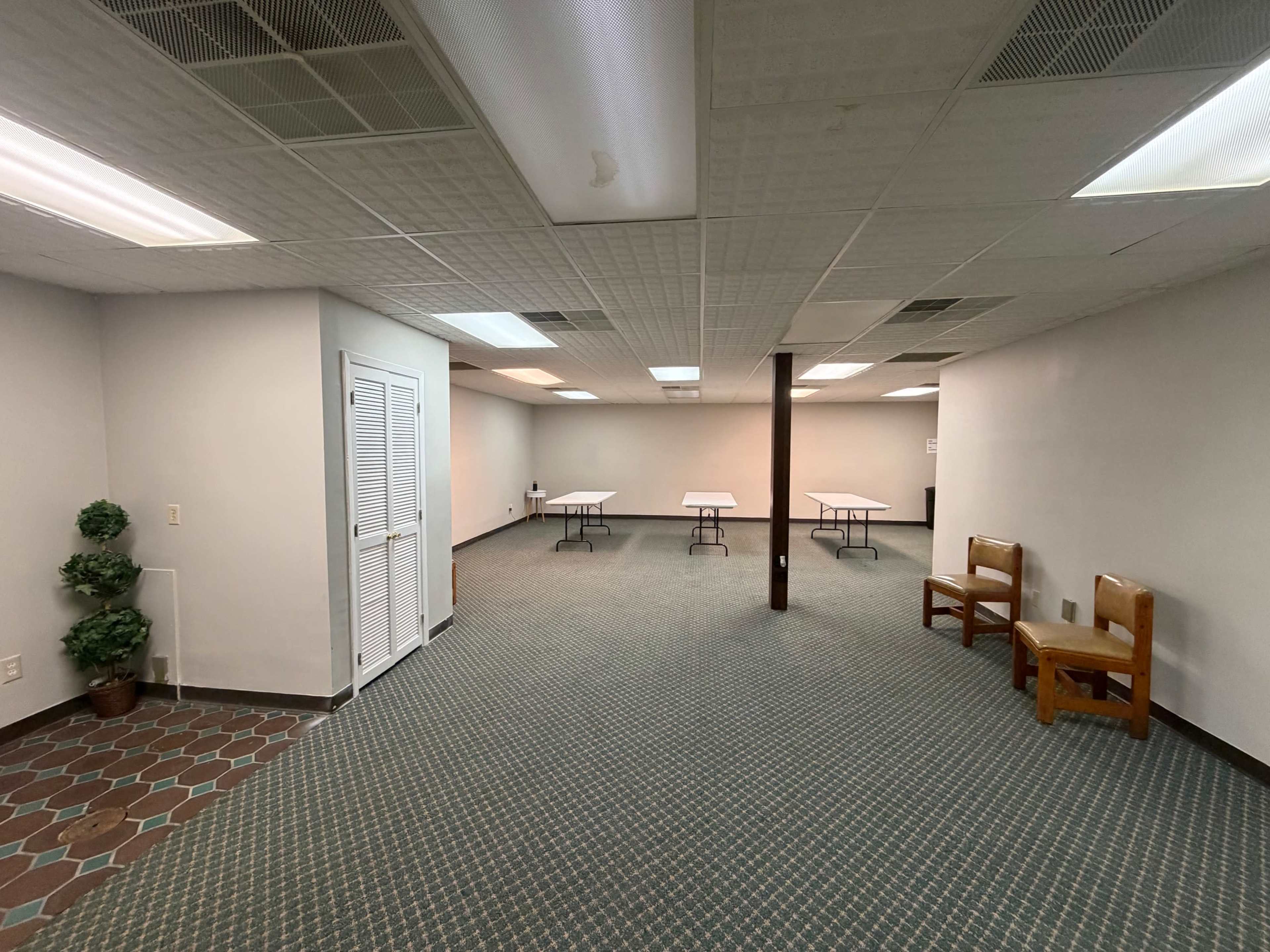 The image shows a spacious, empty room with two wooden chairs, several folding tables, and light-colored walls.