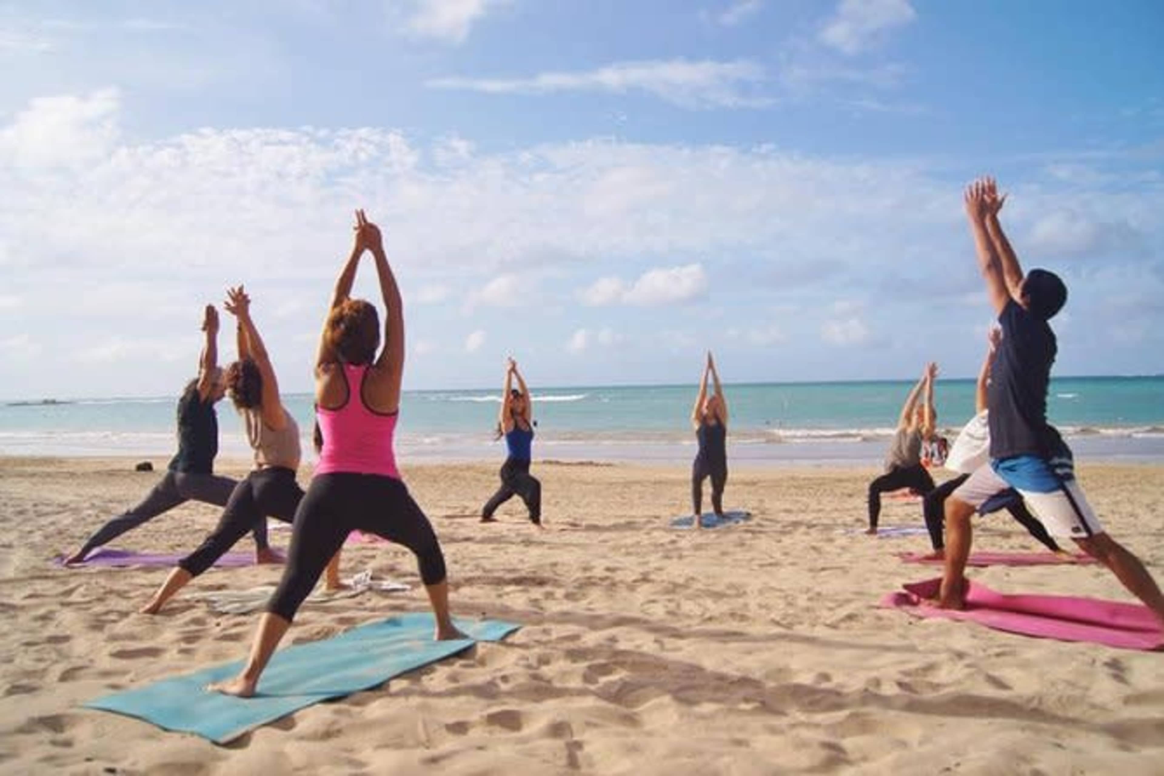 A group of people practices yoga on colorful mats along a sandy beach with the ocean in the background.