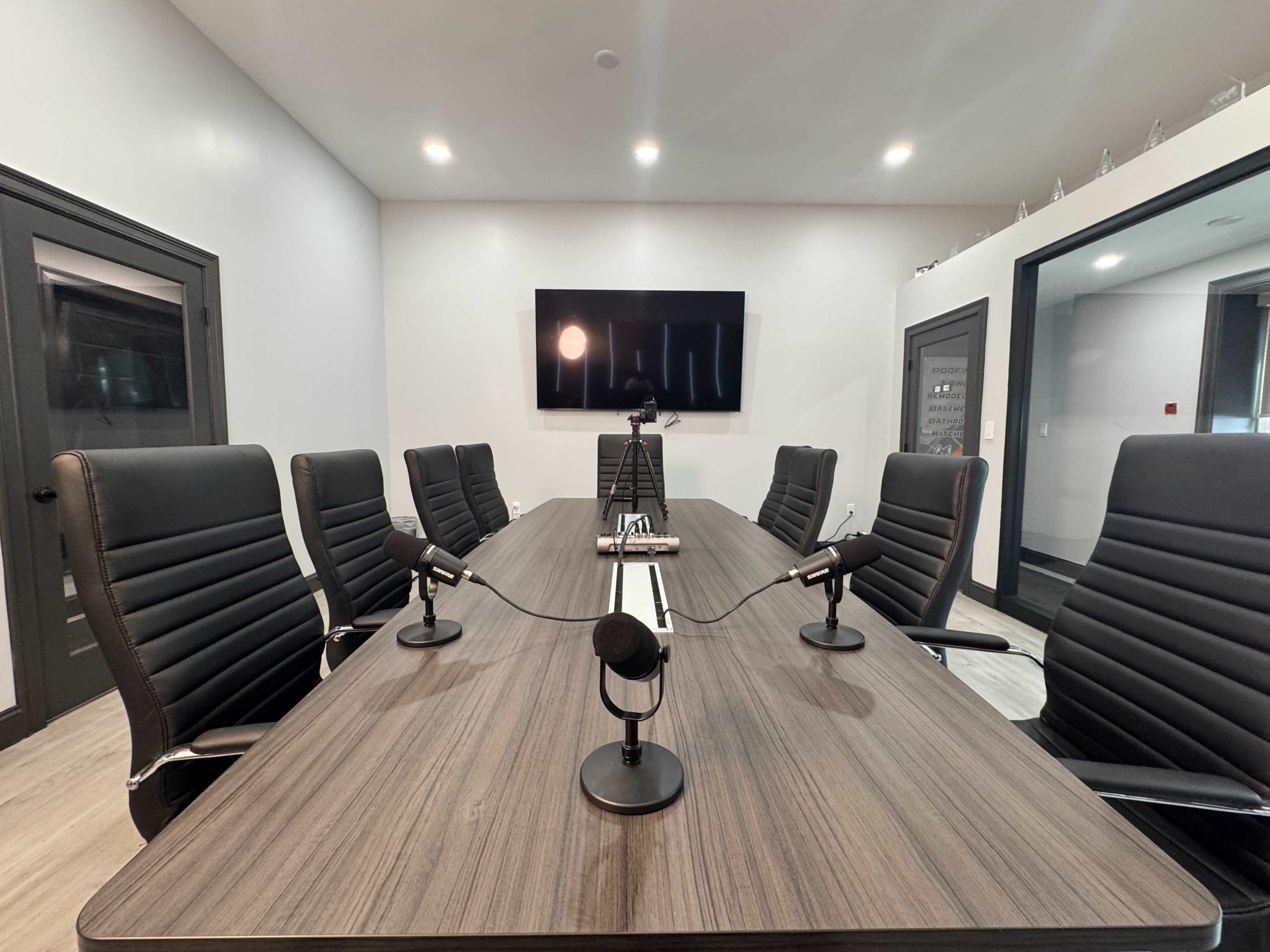 A modern conference room features a long wooden table with six black chairs arranged around it, two microphones on stands, and a large screen mounted on the wall.