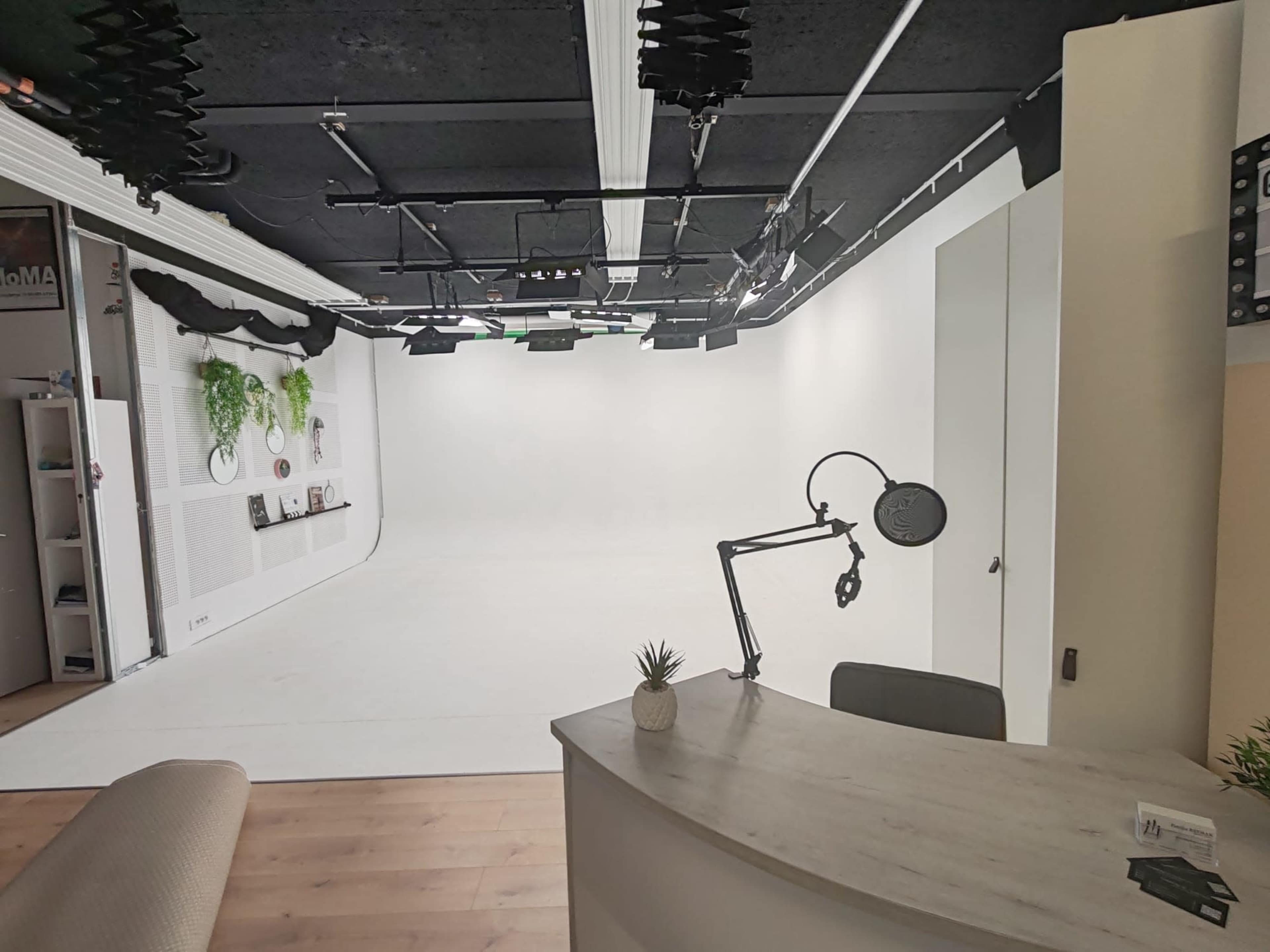 The image shows a bright, empty studio space with a white background, equipped with overhead lighting and a modern reception area featuring a small plant.