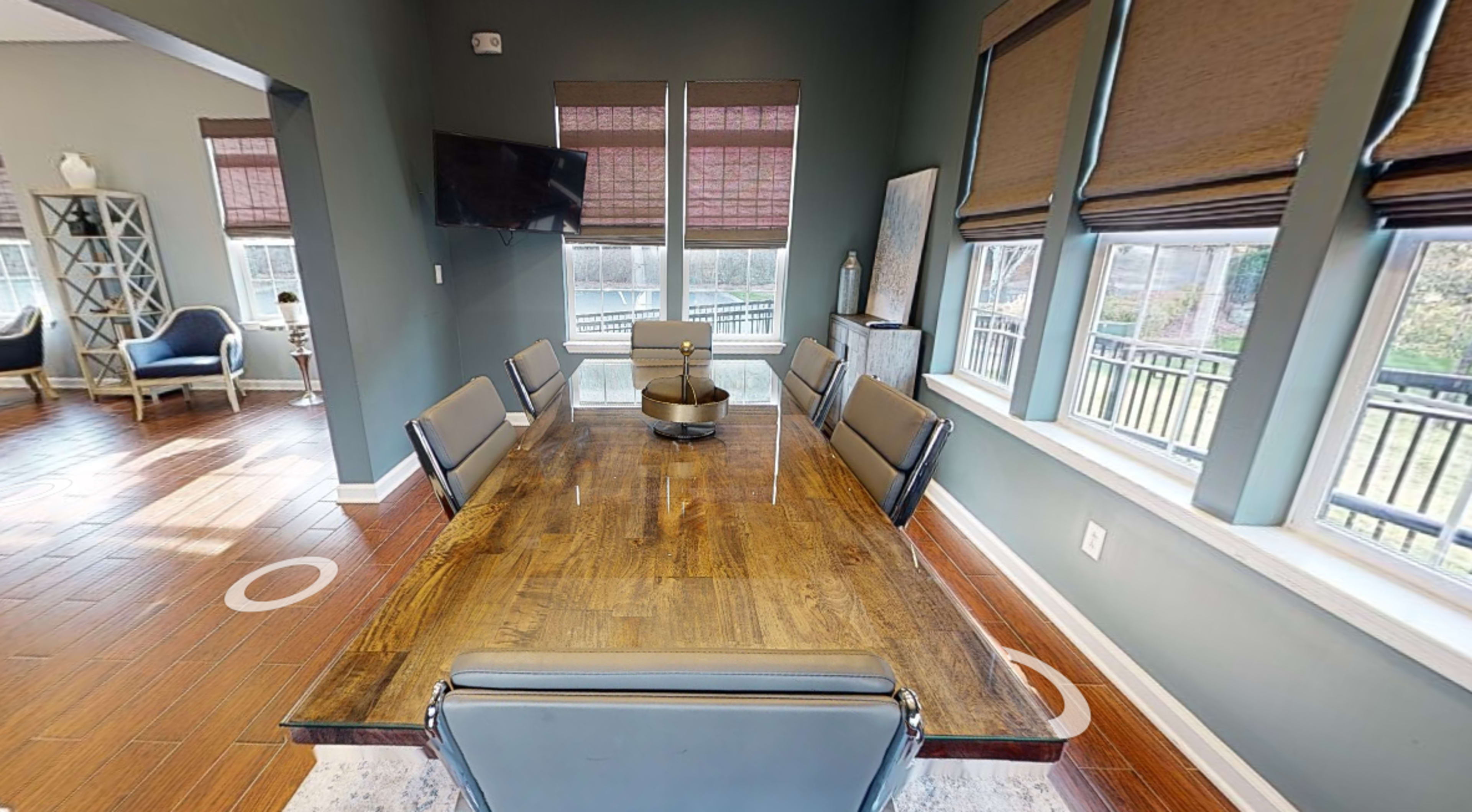 A modern dining area with a long wooden table surrounded by six chairs, large windows with blinds, and a television mounted on the wall.