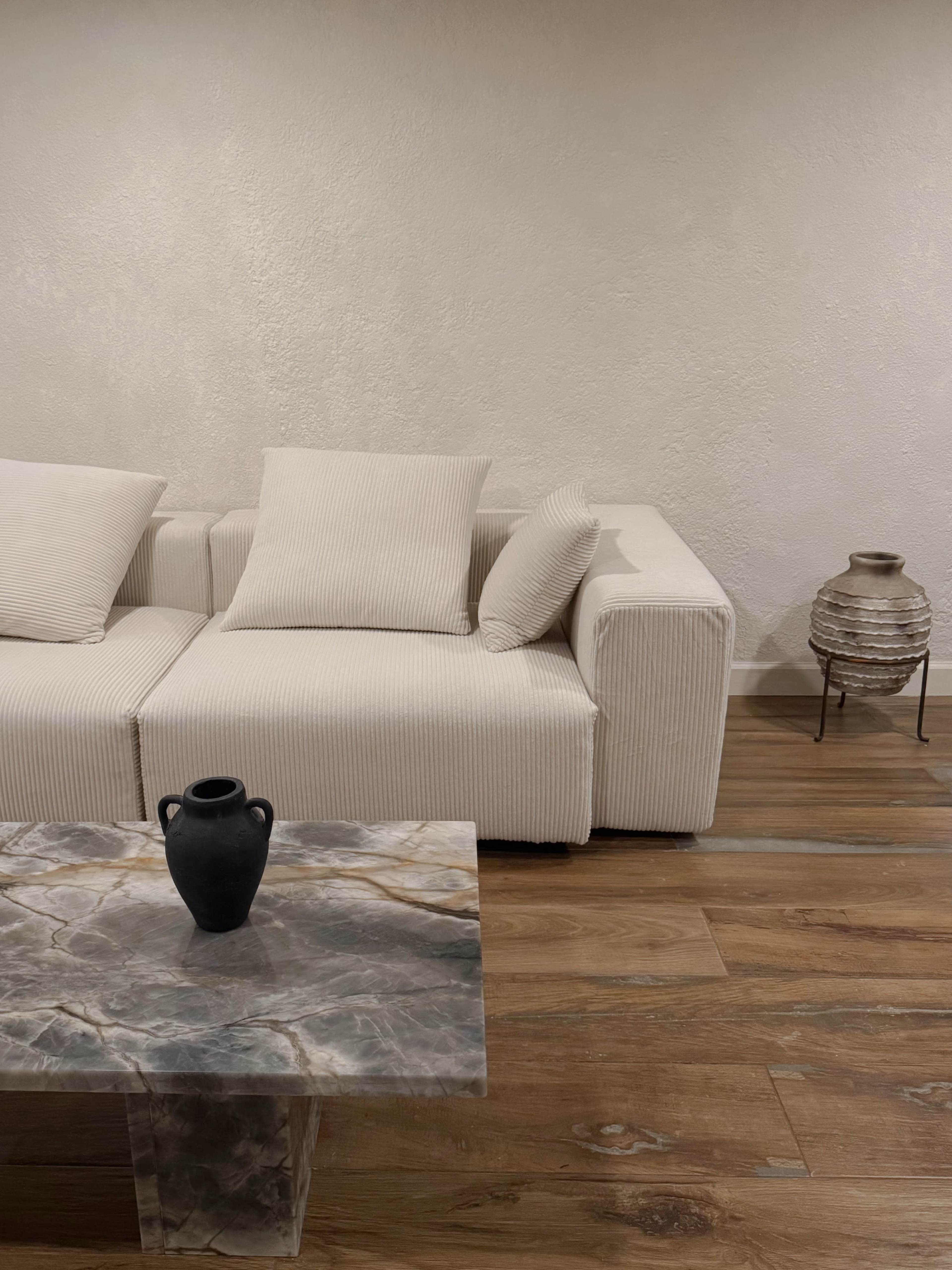 A light-colored sofa with striped cushions is positioned next to a marble coffee table and a decorative vase on the table in a minimalist room.