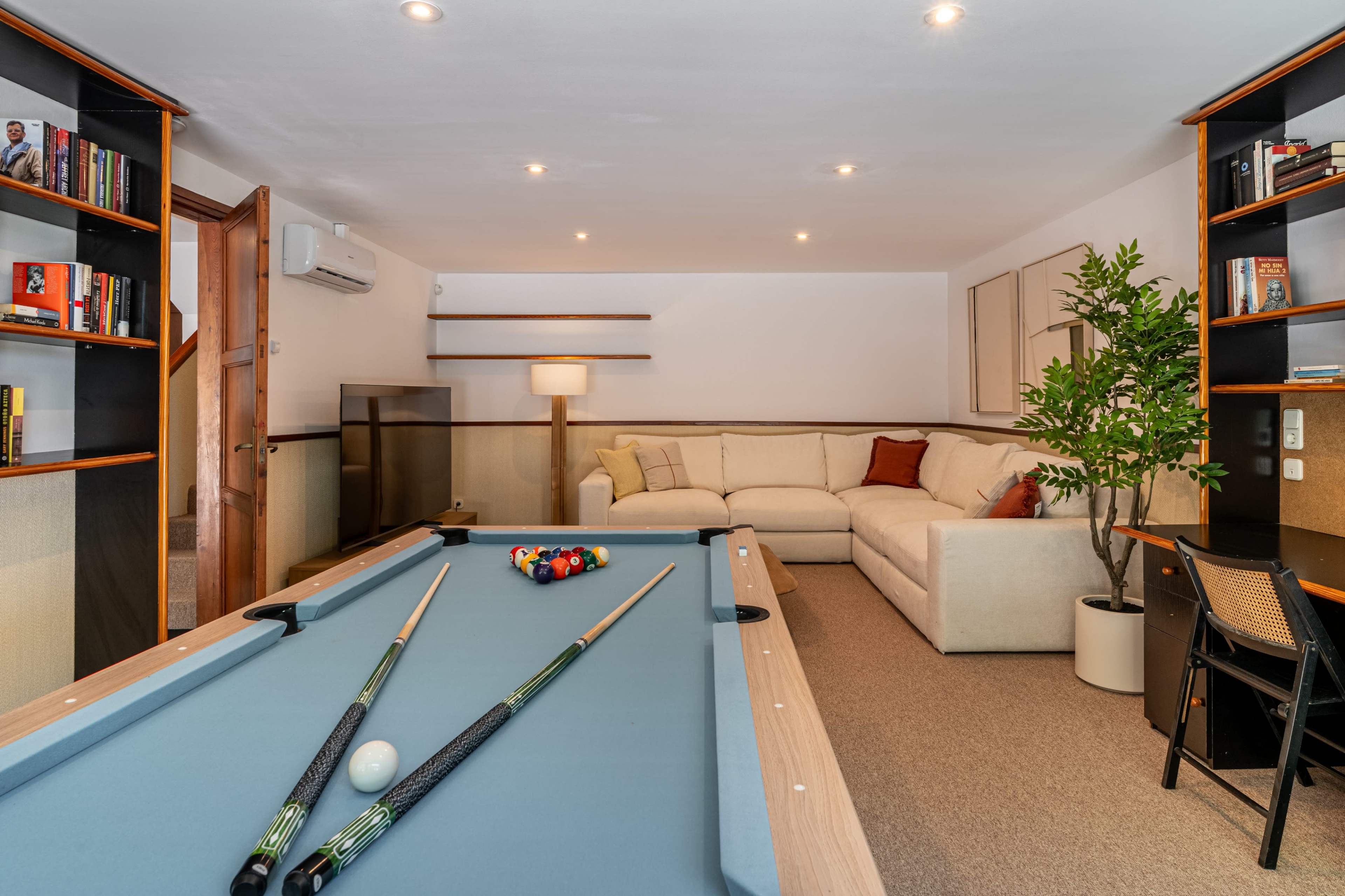 A cozy recreational room features a pool table, a white sectional sofa, bookshelves filled with books, and a small indoor plant.