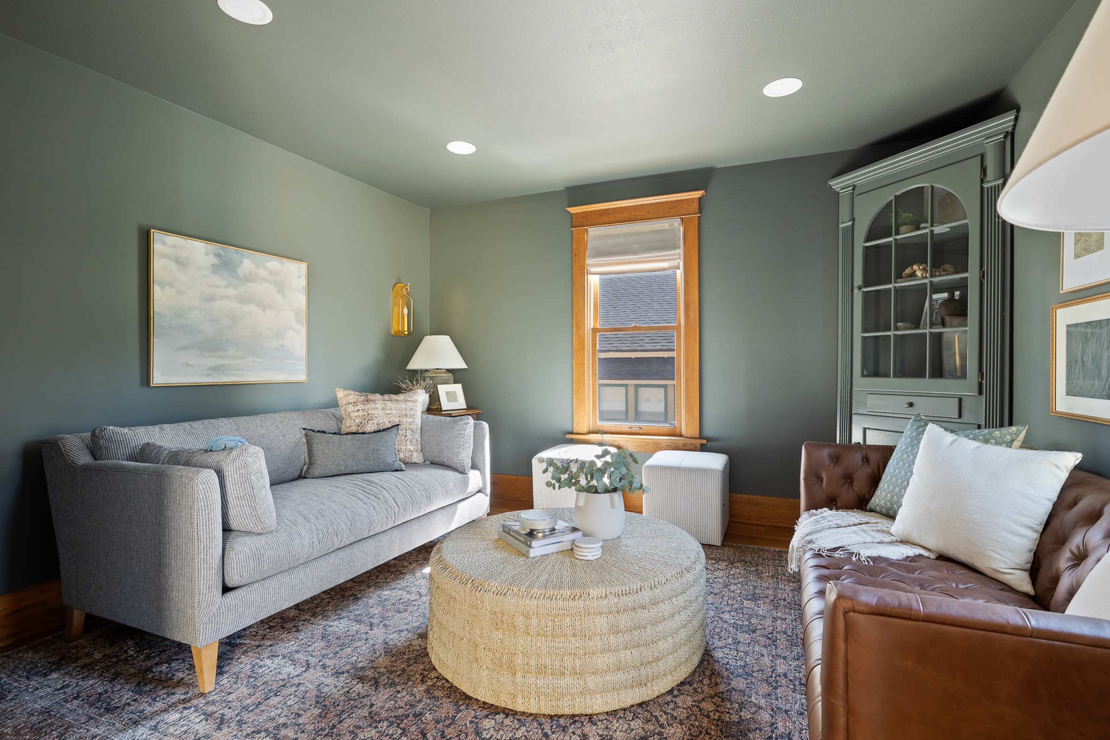 A cozy living room with a gray sofa, a brown leather couch, a round woven coffee table, and a window with natural light.