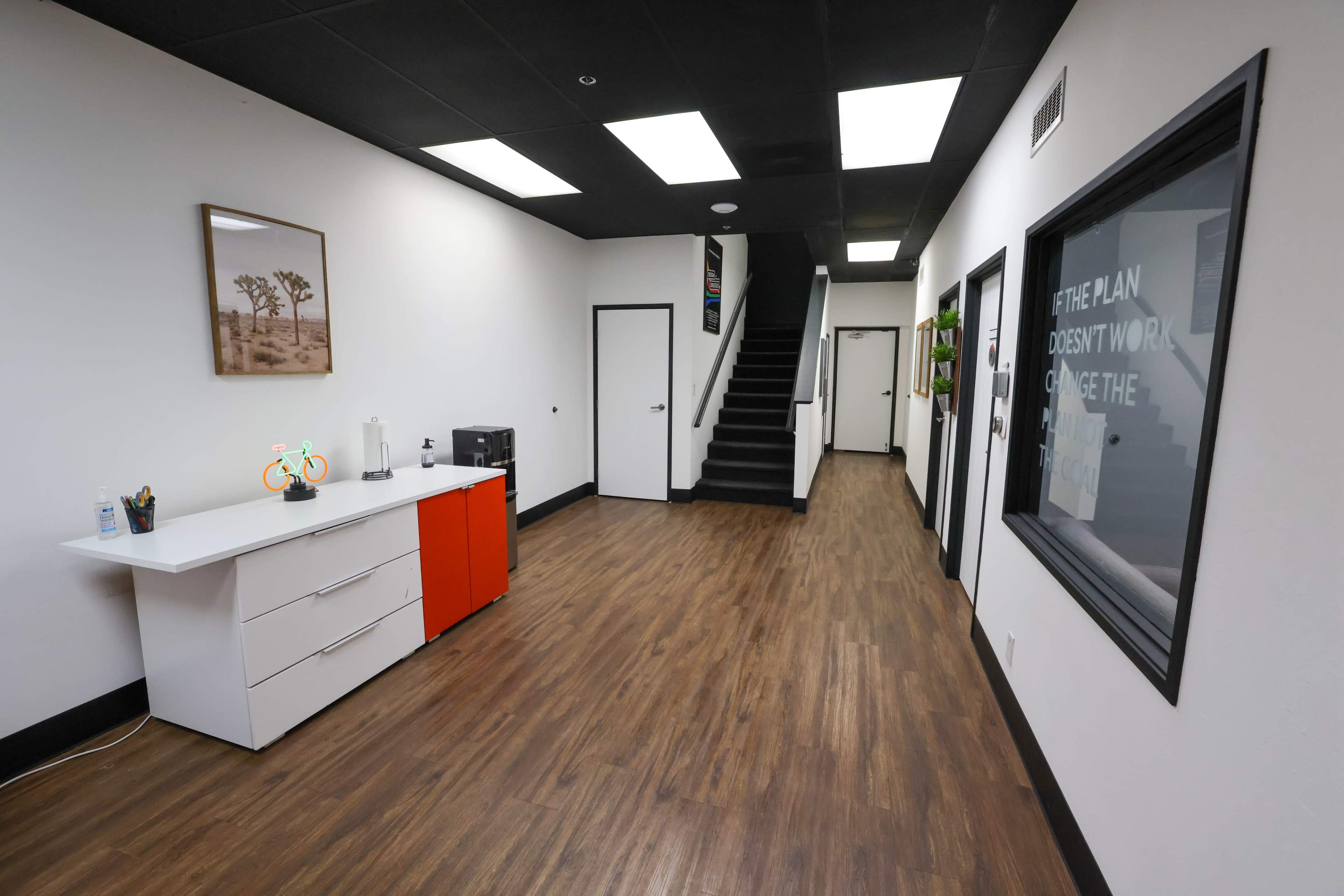 A modern office hallway features a white and orange reception desk, a staircase at the back, and framed artwork on the walls.