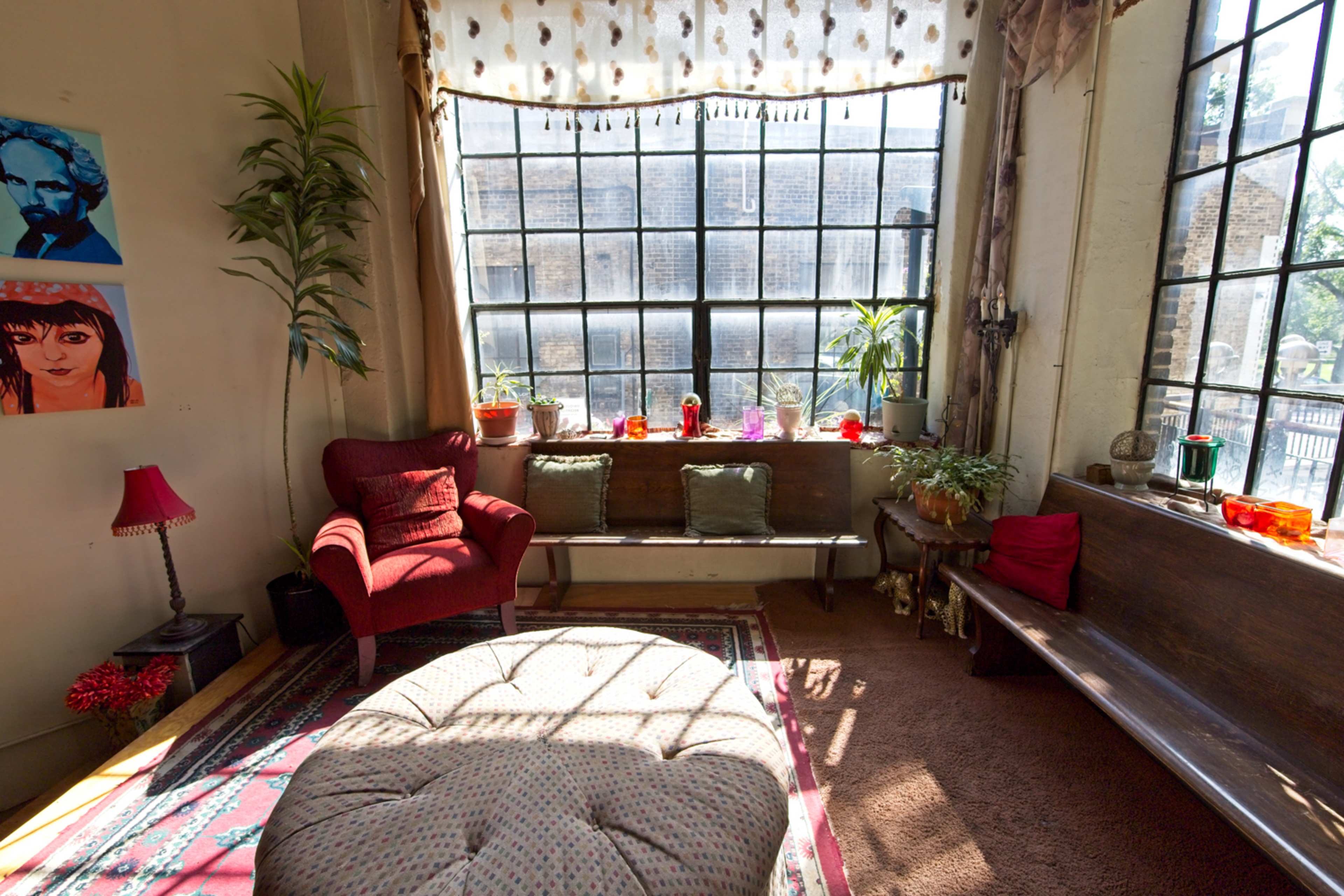 A cozy living room features a large window with plants and colorful decor, including a red chair and a round ottoman.