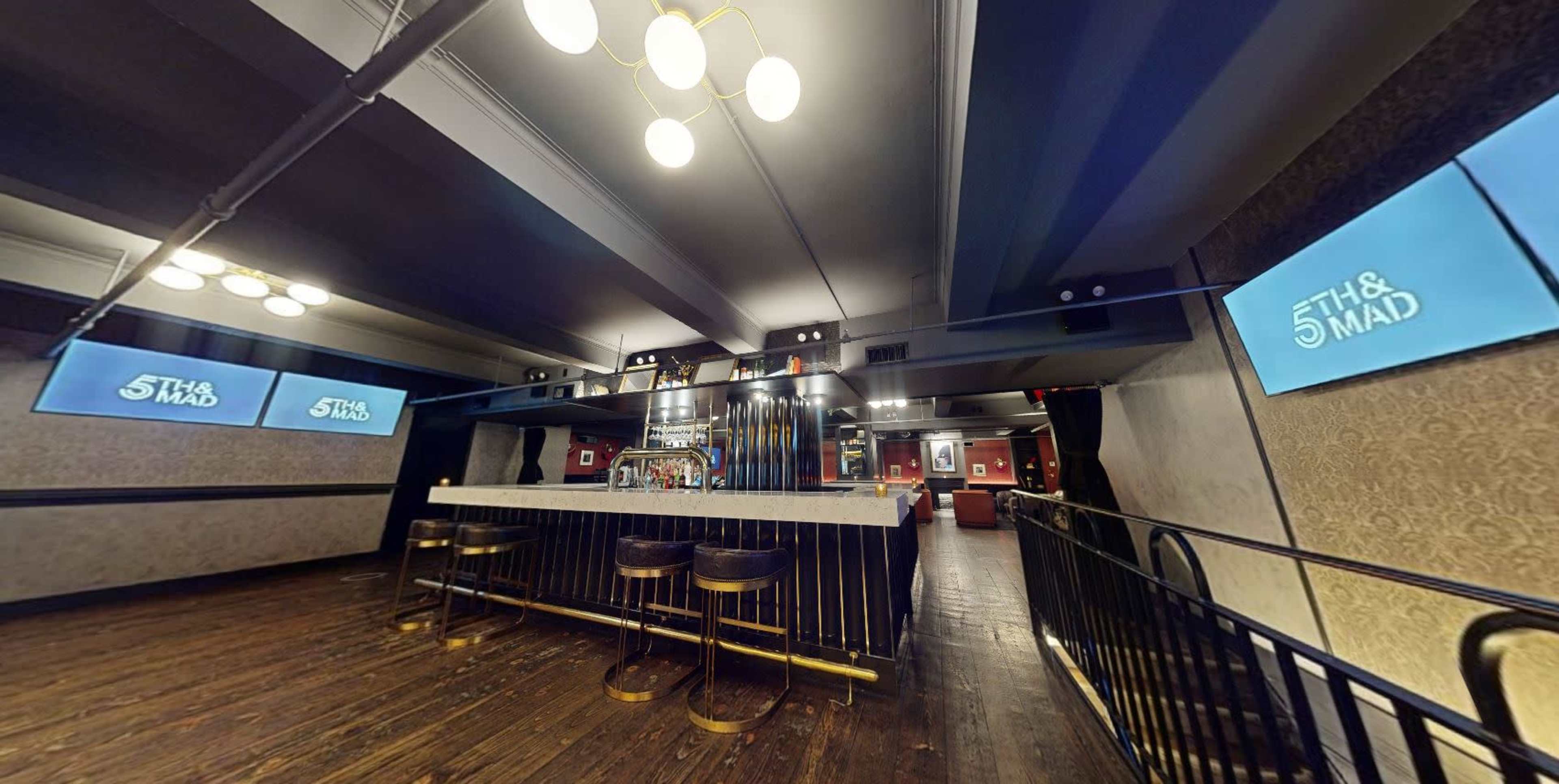 The image shows a well-lit bar area with a sleek countertop, high stools, and digital screens displaying the name "5th & Mad."