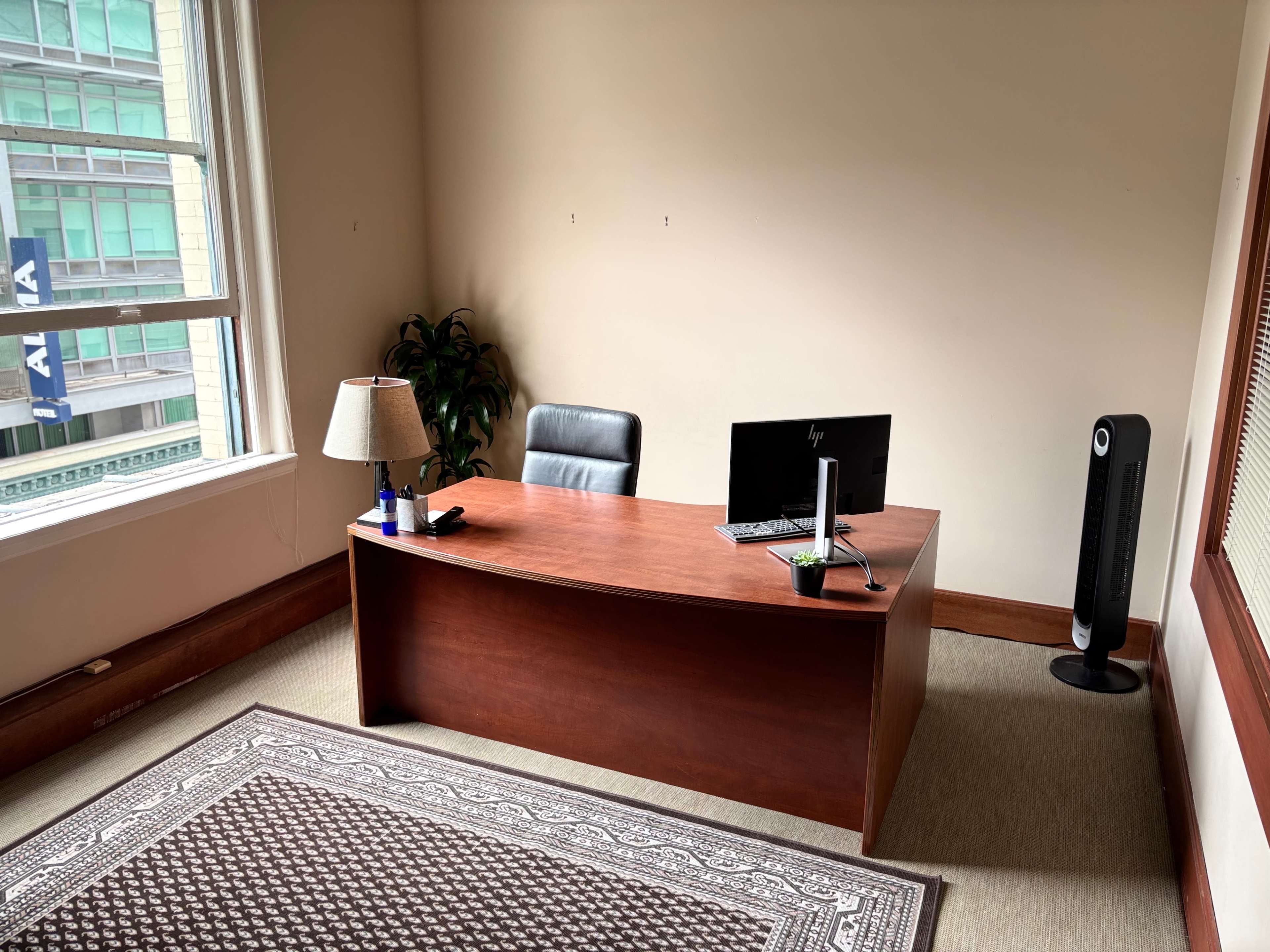 The image shows a simple office space with a wooden desk, a black office chair, a computer monitor, a small lamp, a plant, and a space heater, all against a plain wall and a window with city views.