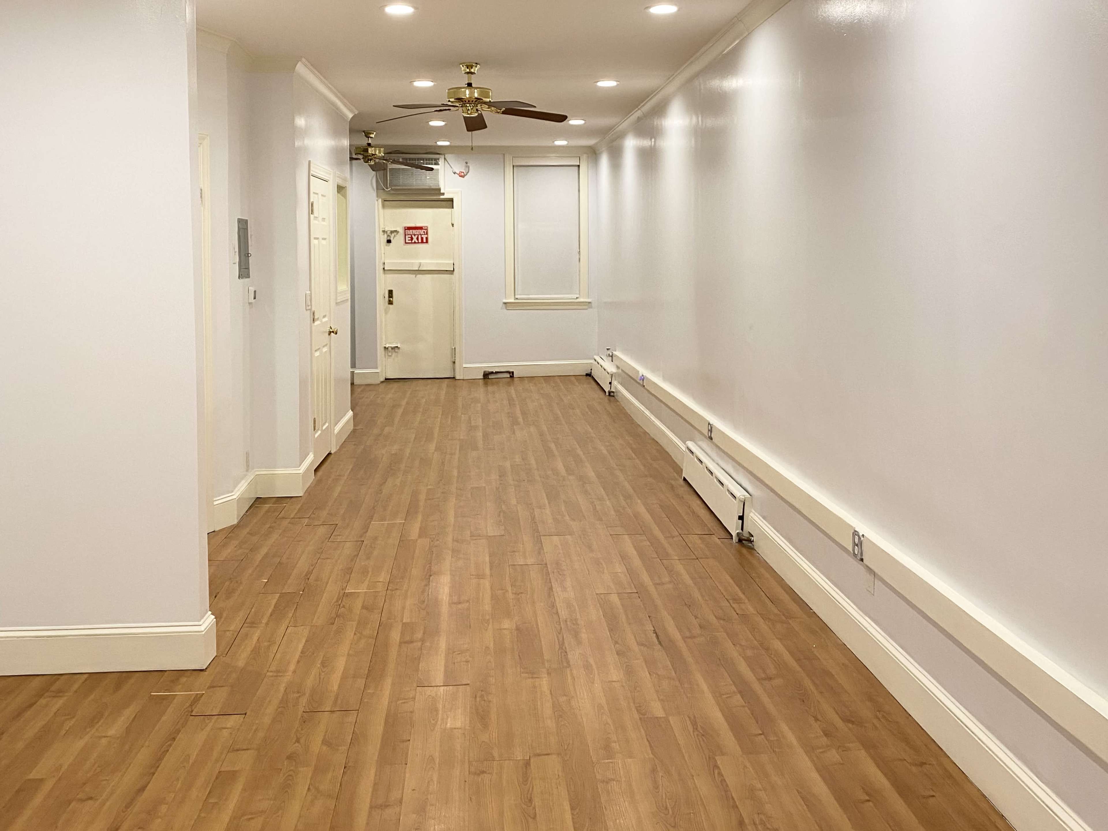 The image shows a long, narrow hallway with wooden flooring and white walls, featuring ceiling lights and a ceiling fan.