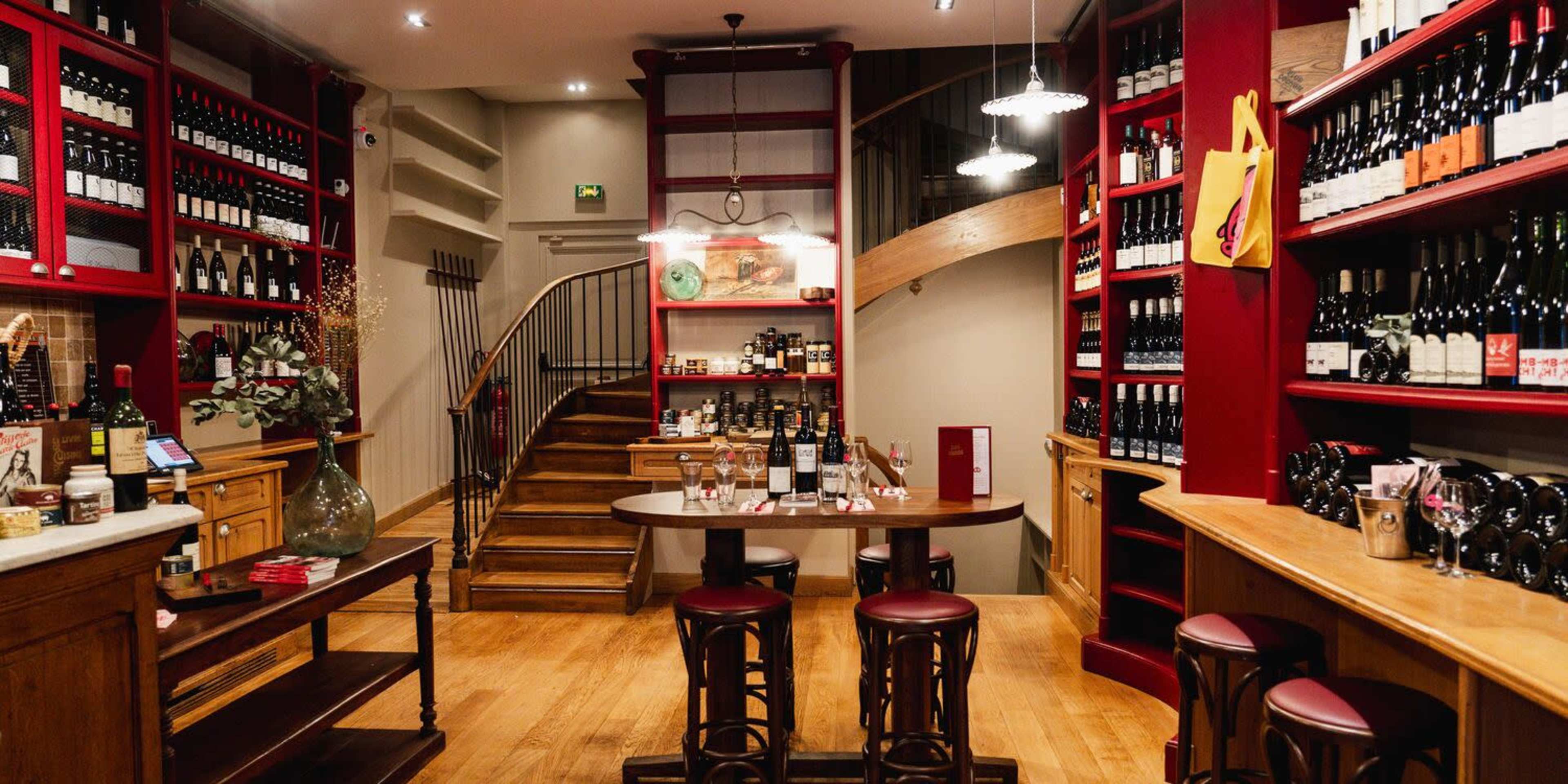 A wine shop features wooden shelves stocked with various bottles, a central tasting table with stools, and a staircase leading to an upper level.