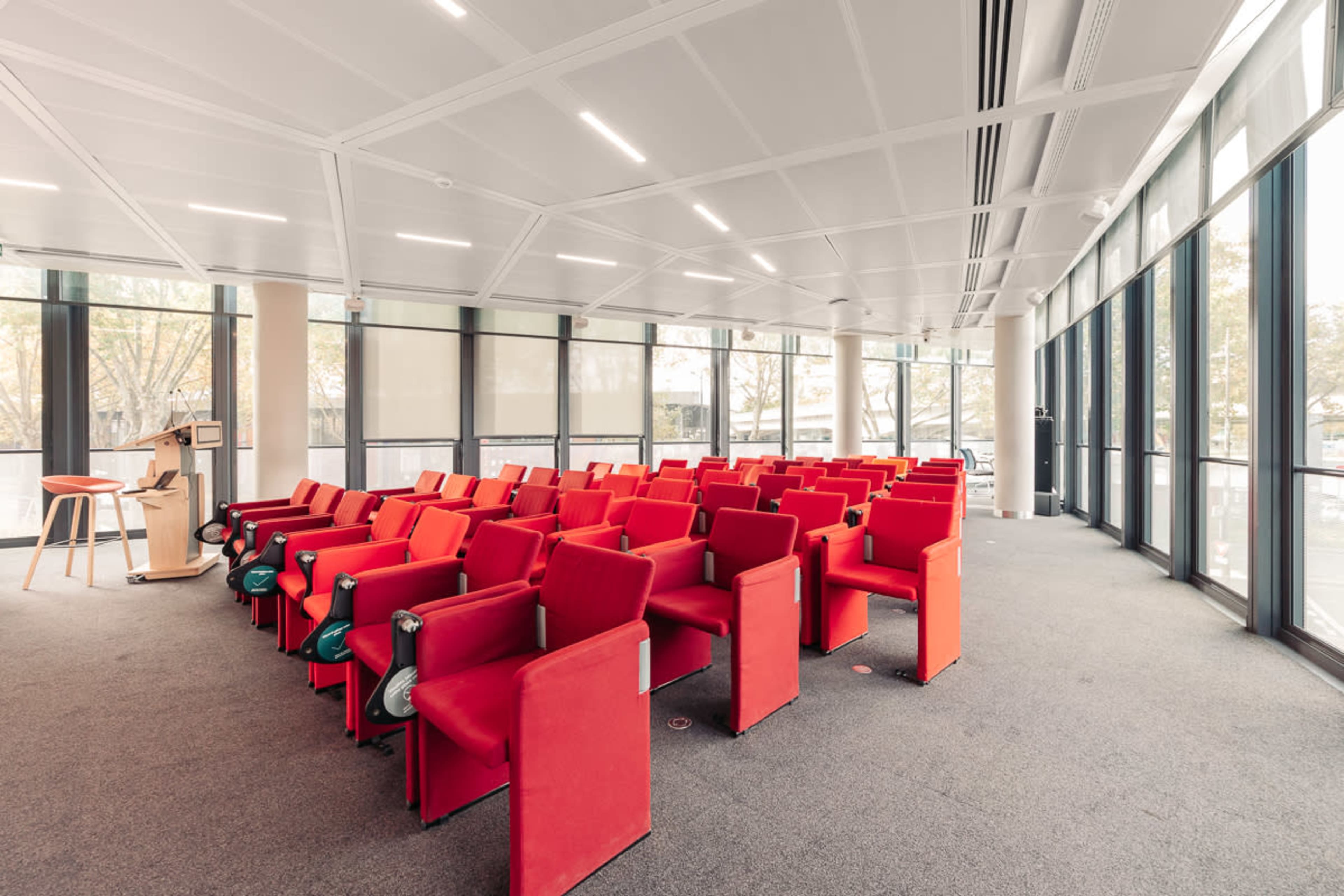 The image shows a modern conference room featuring rows of red chairs arranged in a semi-circle, with large windows letting in natural light.