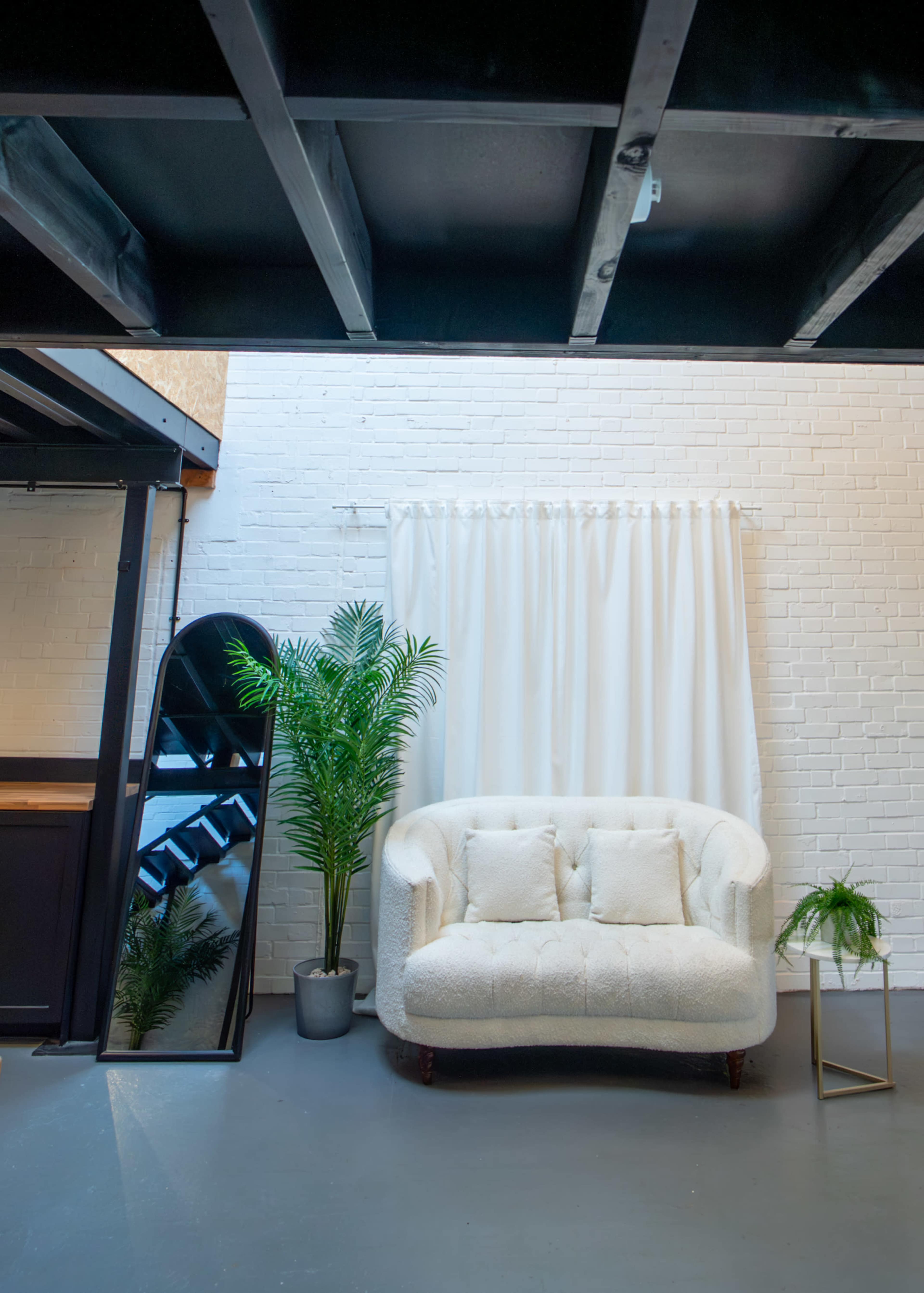 The image features a cozy living area with a white tufted couch, a large mirror, a potted plant, and sheer curtains against a white brick wall.